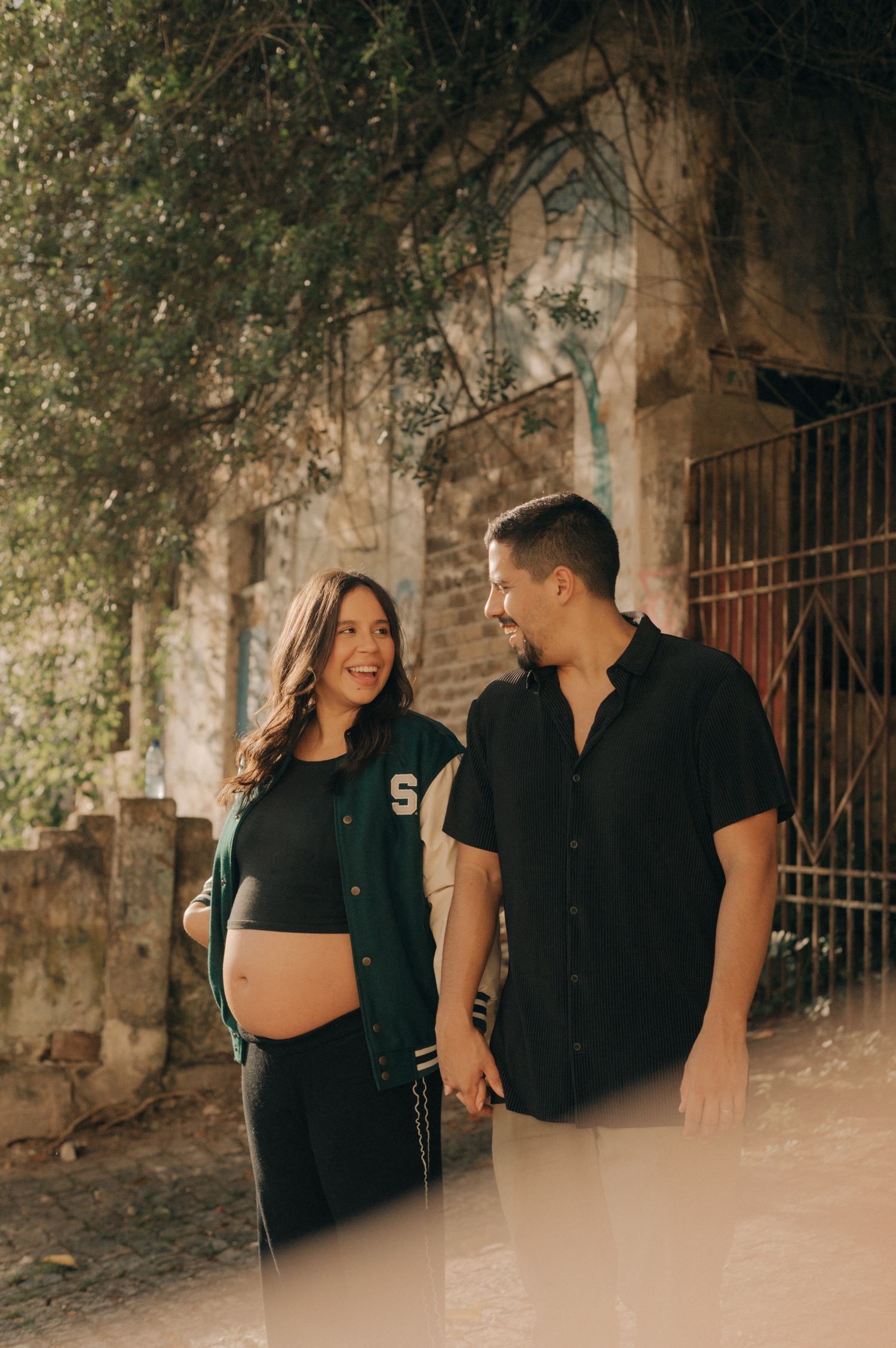 Expectant couple in casual black outfits pose together against a rustic stone wall.