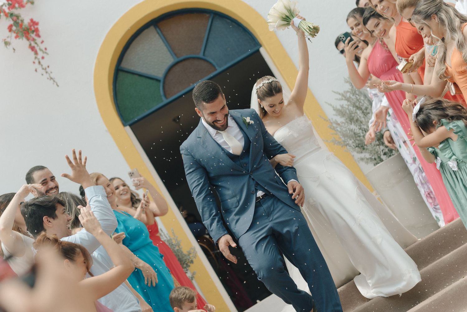 Wedding couple exits through an archway with colorful stained glass as guests throw confetti.