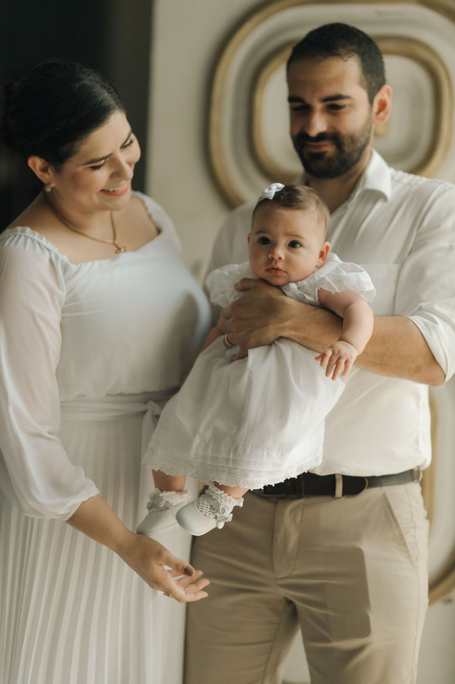 A baby in a white christening dress is held lovingly by parents wearing coordinating white and beige outfits.