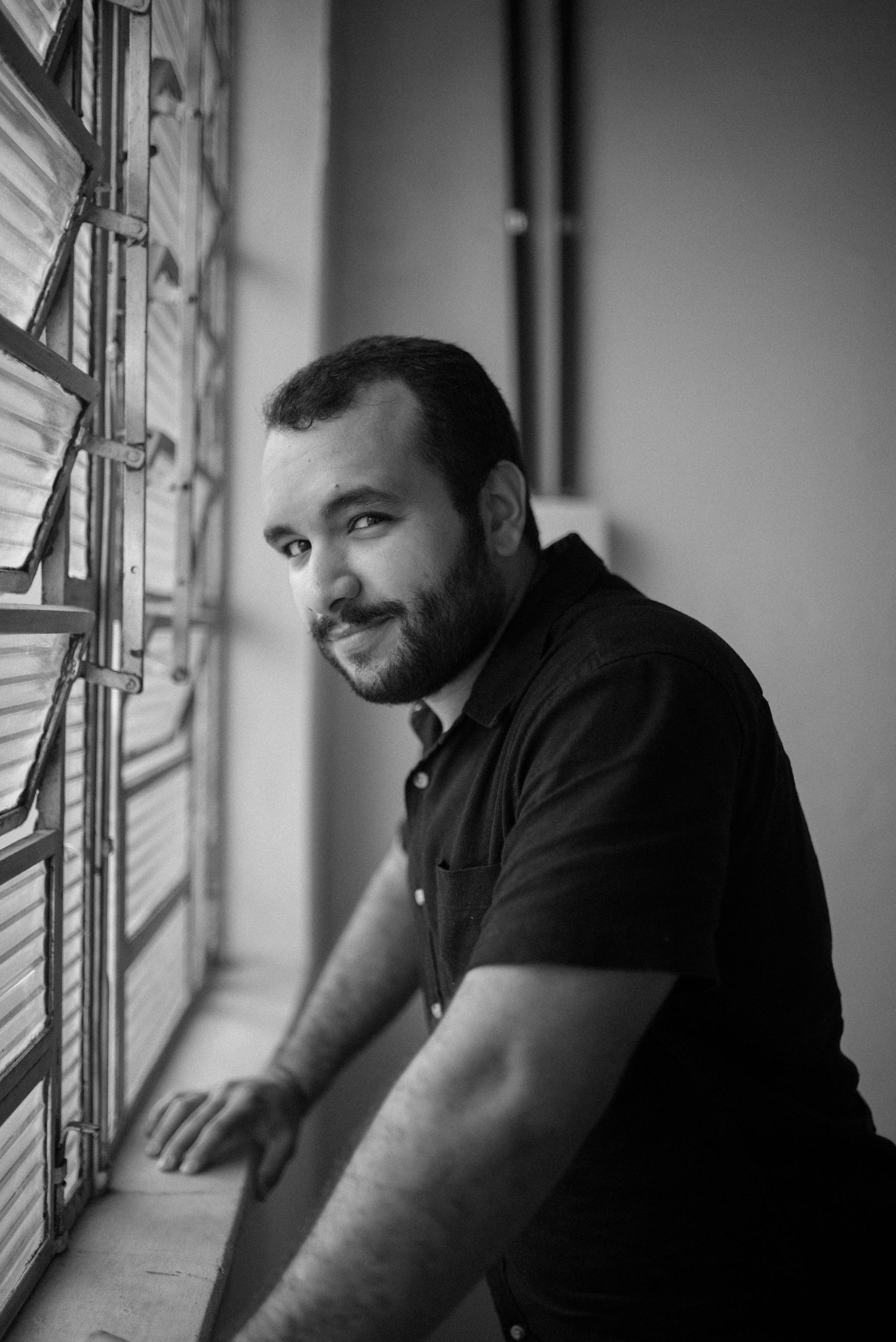 Black and white portrait of man in dark shirt smiling by window.