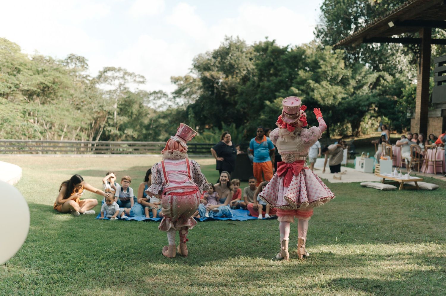 Performers in pink costumes dance on a grassy lawn with guests relaxing in the background.
