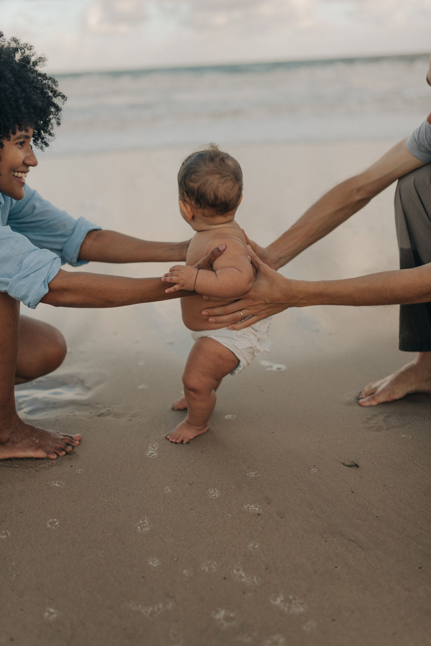 Parents helping their baby take first steps on beach at sunset.