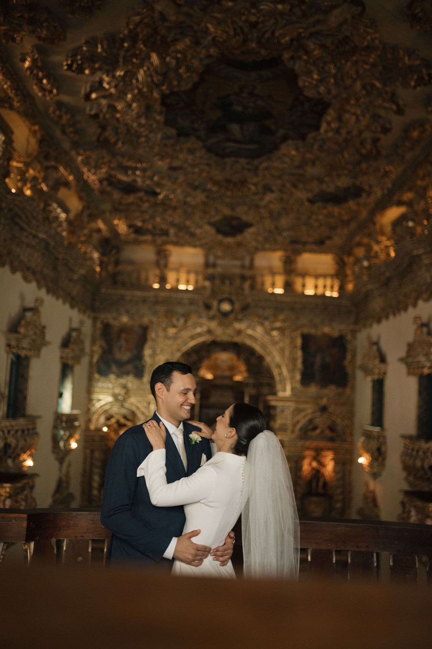 Bride and groom embrace in ornate golden baroque church interior during wedding ceremony.