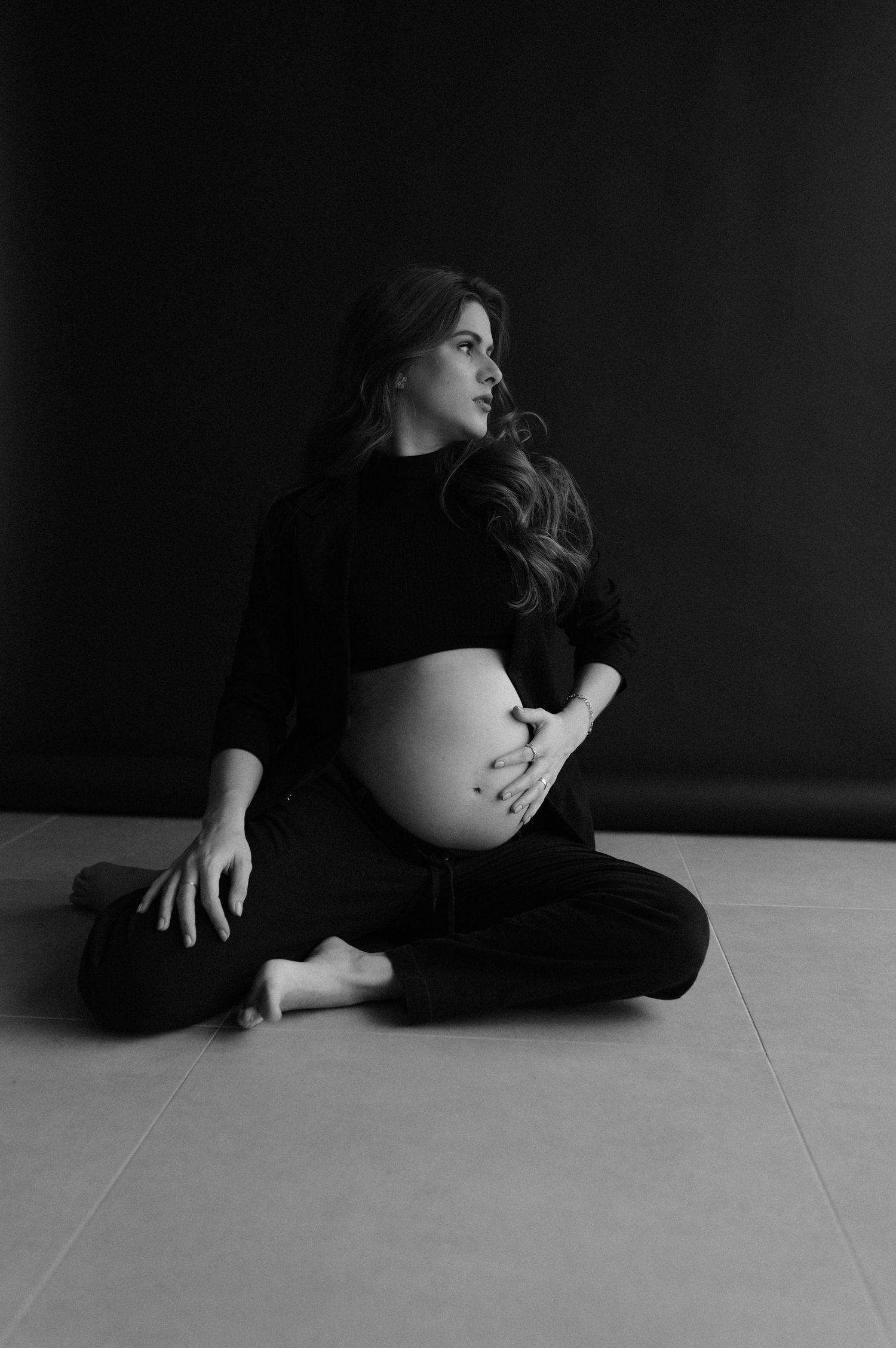 Artistic black and white maternity photo of expecting mother sitting cross-legged in dark clothing against dark background.