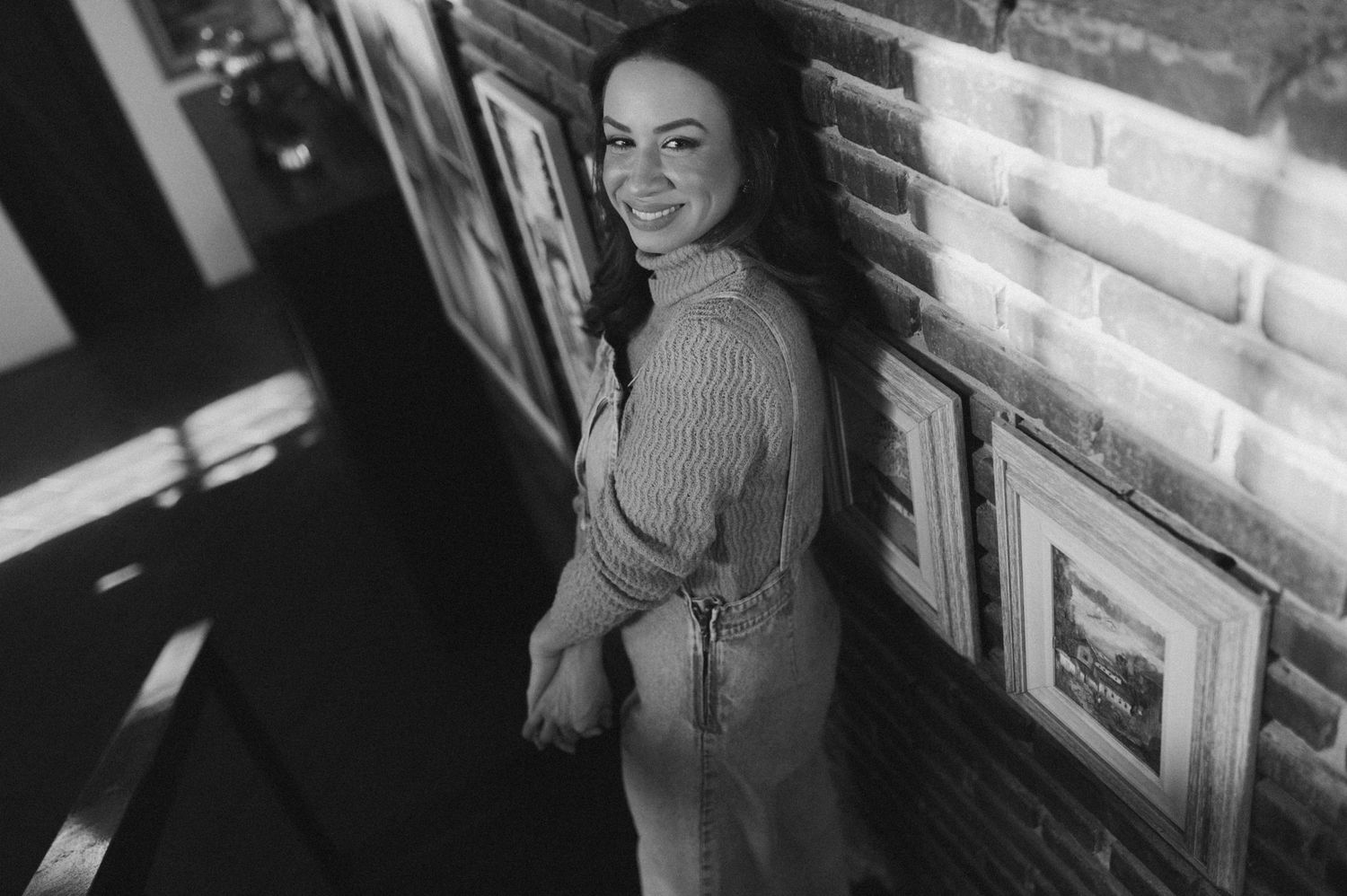 Black and white candid shot of someone posing by a brick wall with framed artwork.