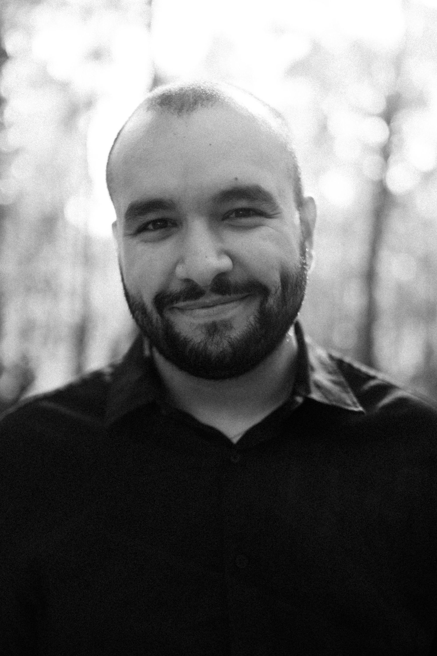 Black and white portrait photo showing a bearded man in a dark shirt smiling warmly against a blurred outdoor background.