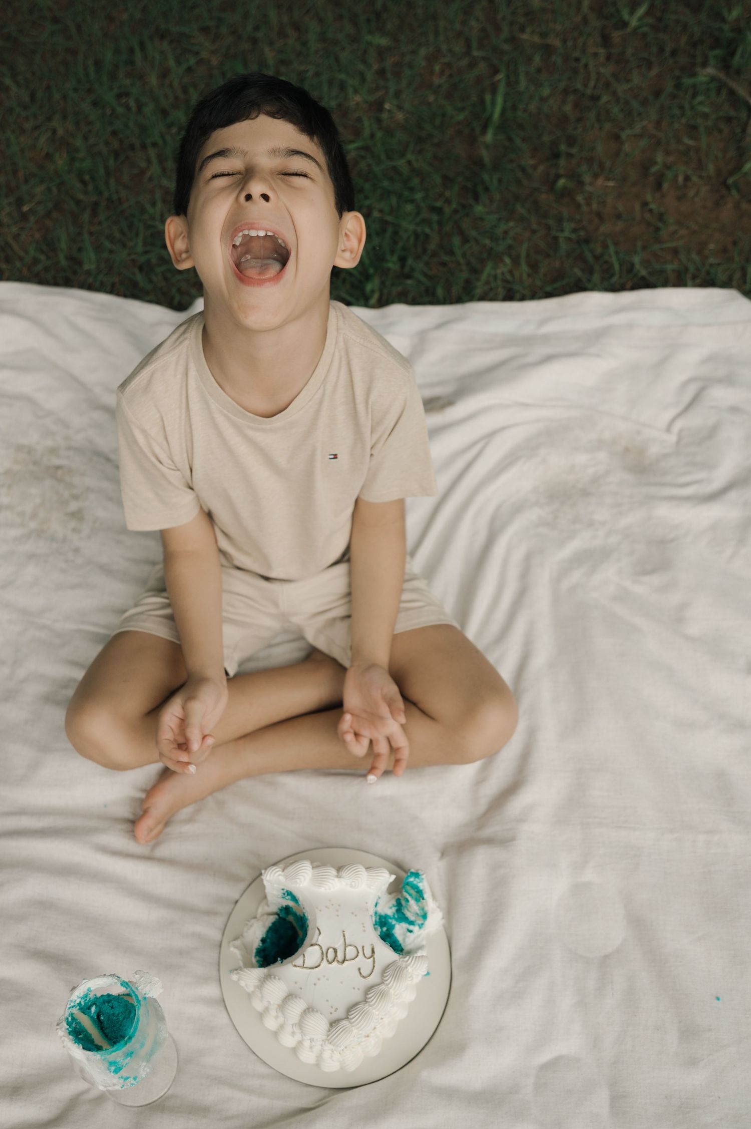 A young boy sits cross-legged on a white blanket outdoors, mouth open wide, next to a white cake labeled Baby.