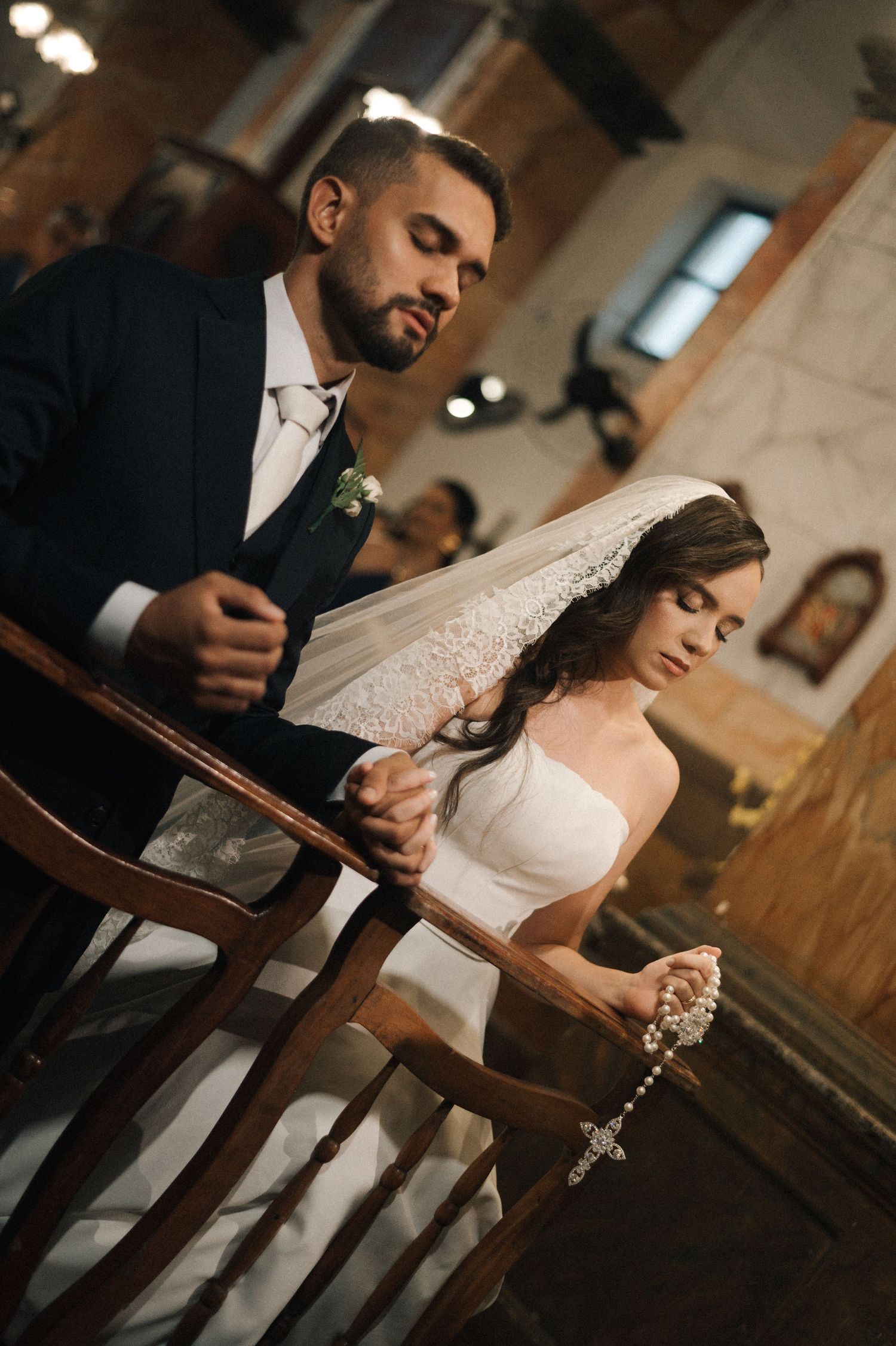 Bride and groom kneeling in prayer at church altar during wedding ceremony, holding a rosary.