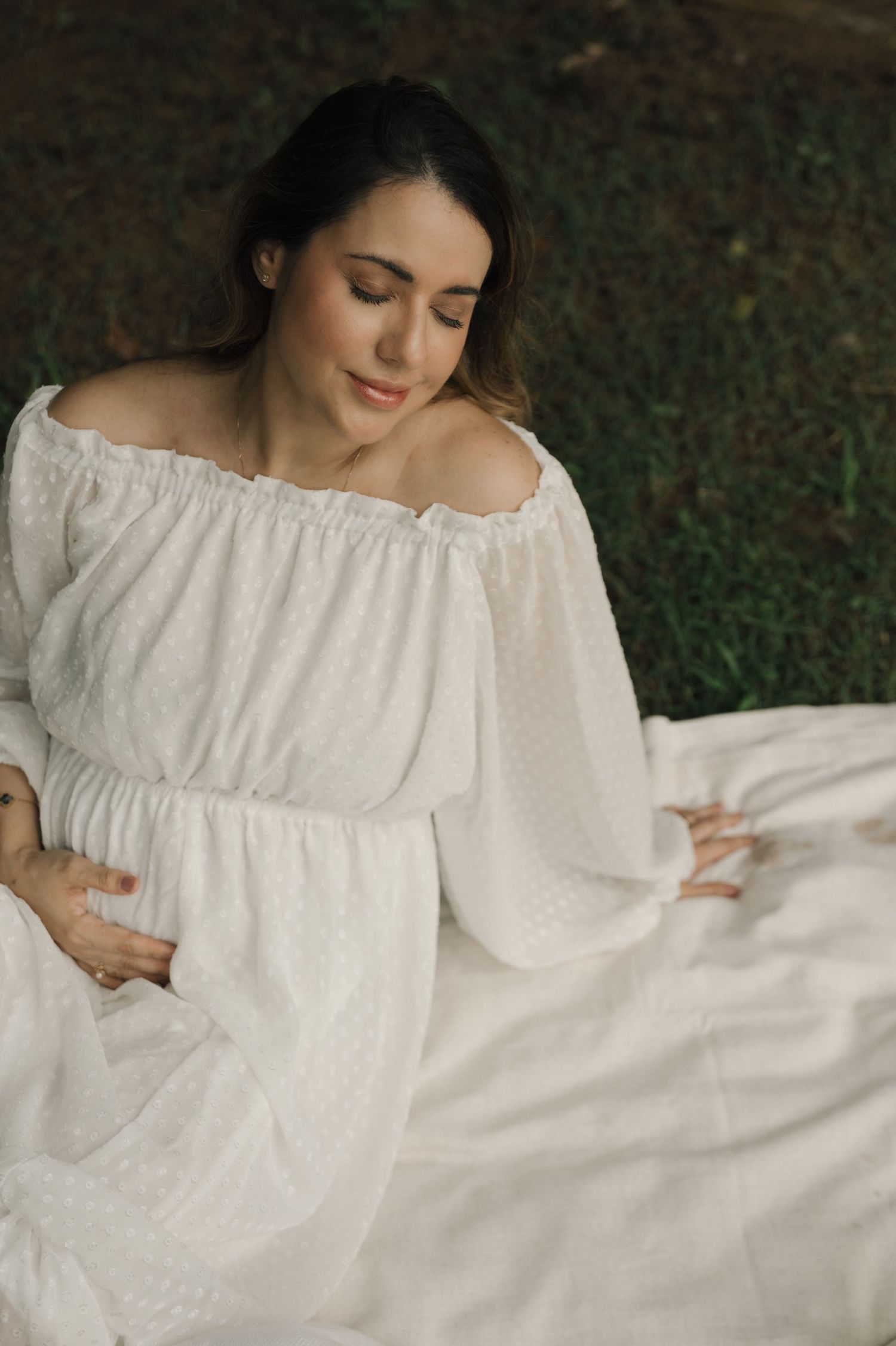 Pregnant woman in white off-shoulder dress sitting outdoors on white fabric, cradling her baby bump with eyes closed.