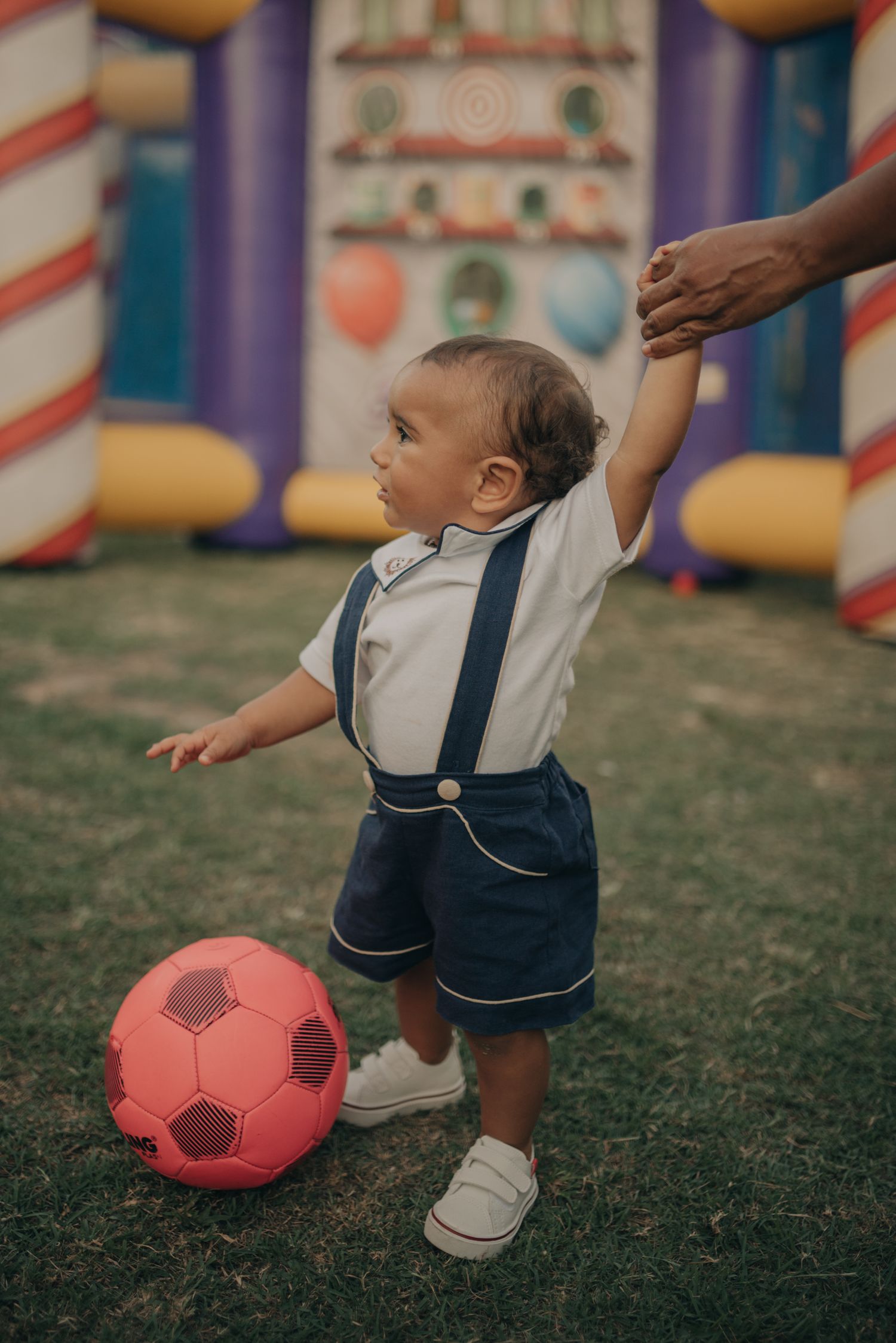 Small child in suspenders playing with red soccer ball near decorative carnival displays.