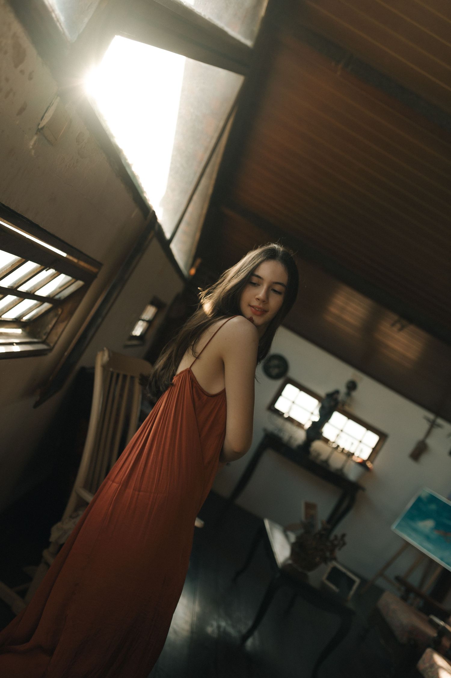 A figure in a flowing rust-colored dress poses in a room with dramatic lighting and wooden beams.