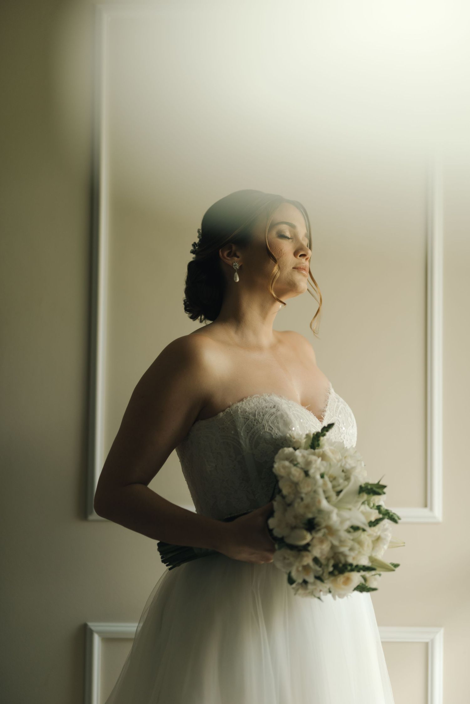 A bride in a strapless gown holds a white bouquet while posing by a window.