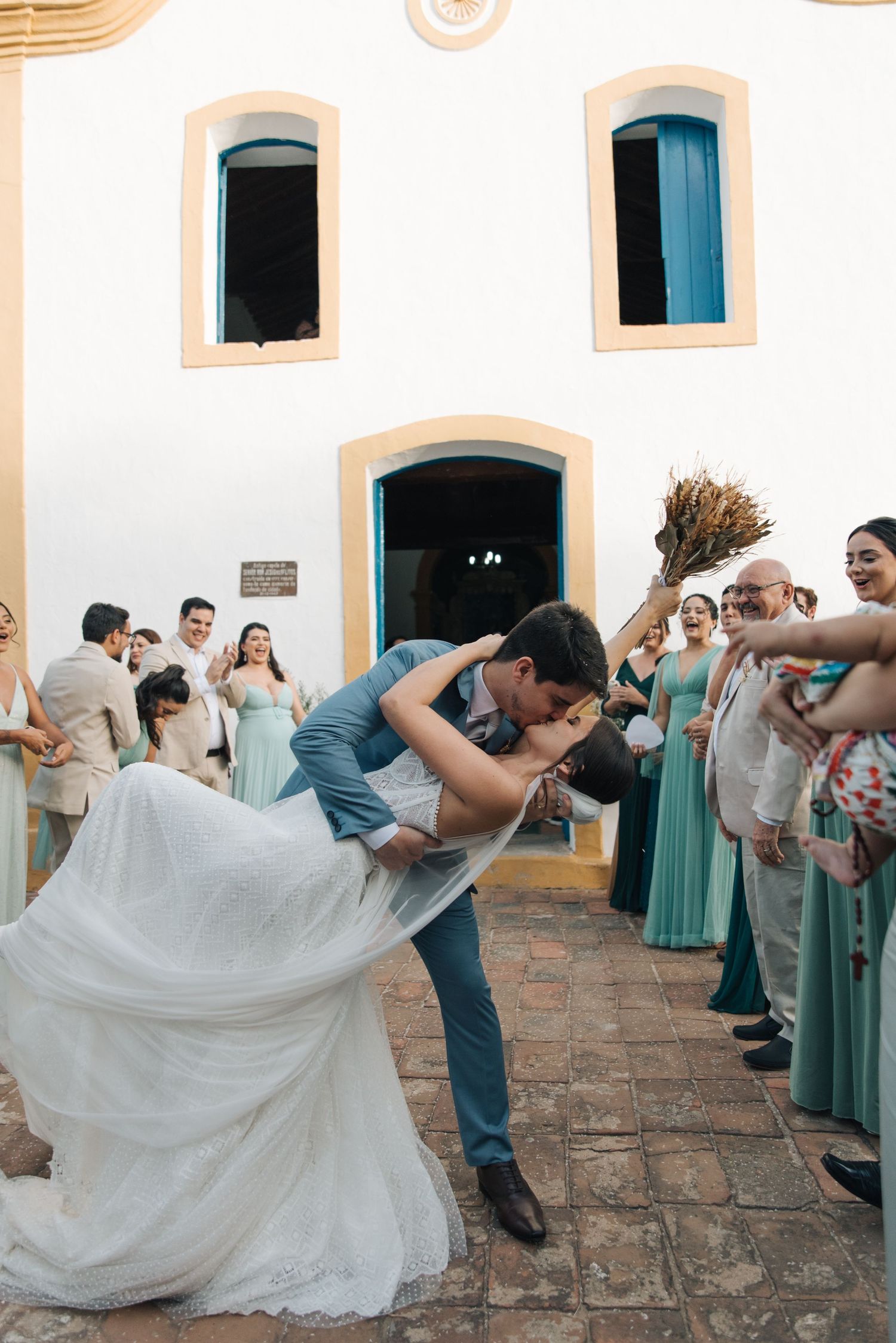 Newlyweds share a romantic dip kiss surrounded by guests in front of a white church.