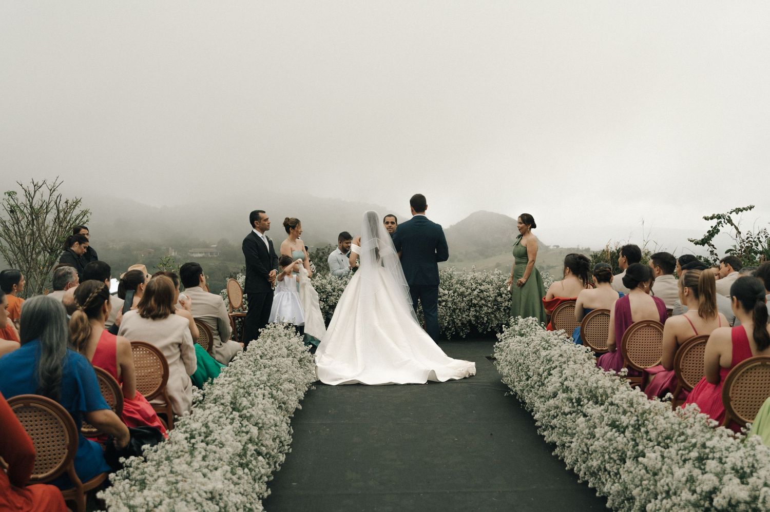 Outdoor wedding ceremony on a misty mountain with white floral aisle decorations and seated guests in coral dresses.