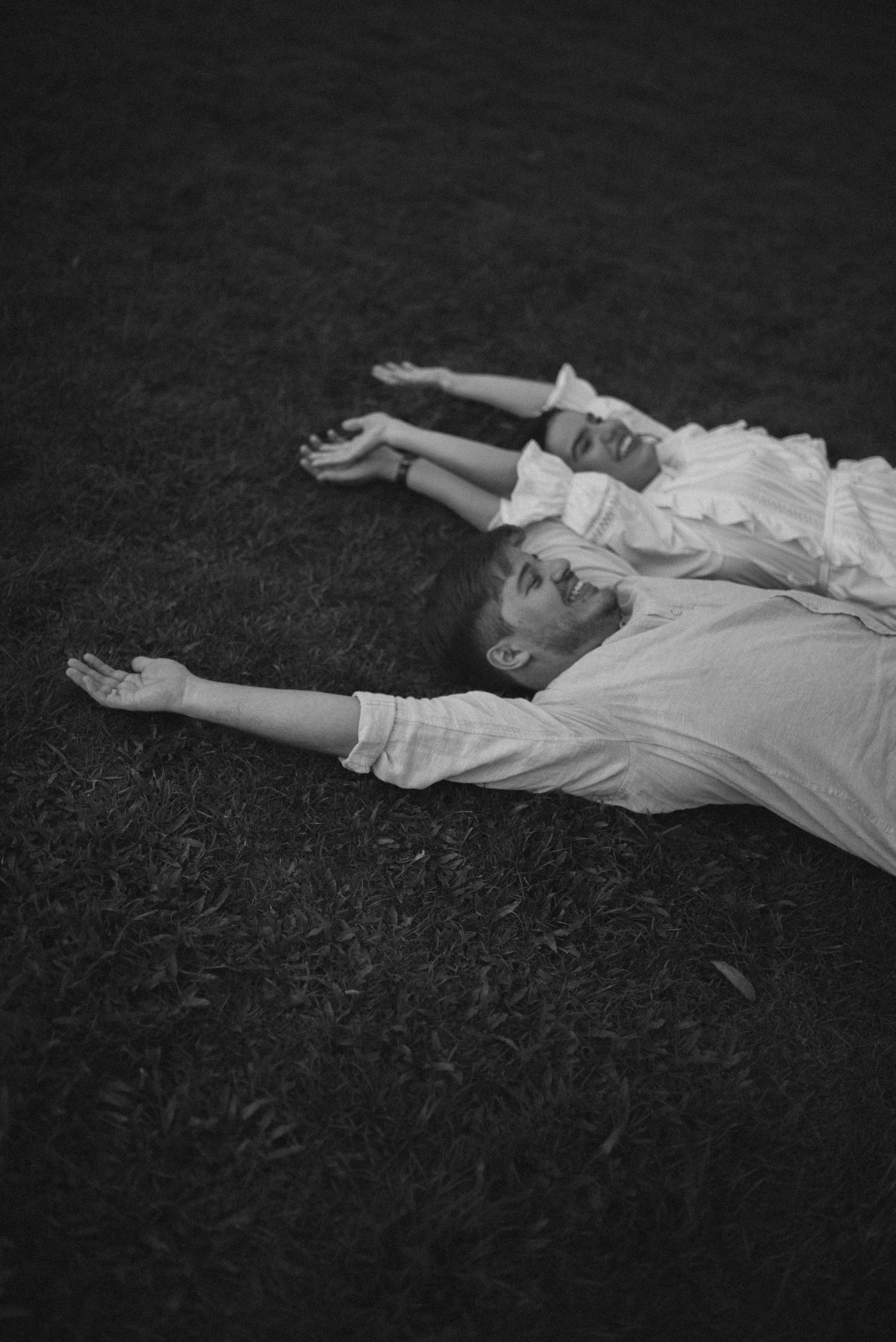 Black and white photo of couple lying on grass with arms outstretched.
