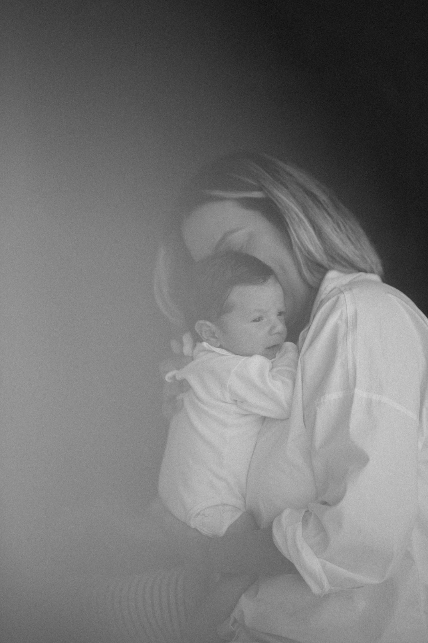 A sleeping baby lies peacefully in a white onesie against a soft white background in a monochrome photograph.