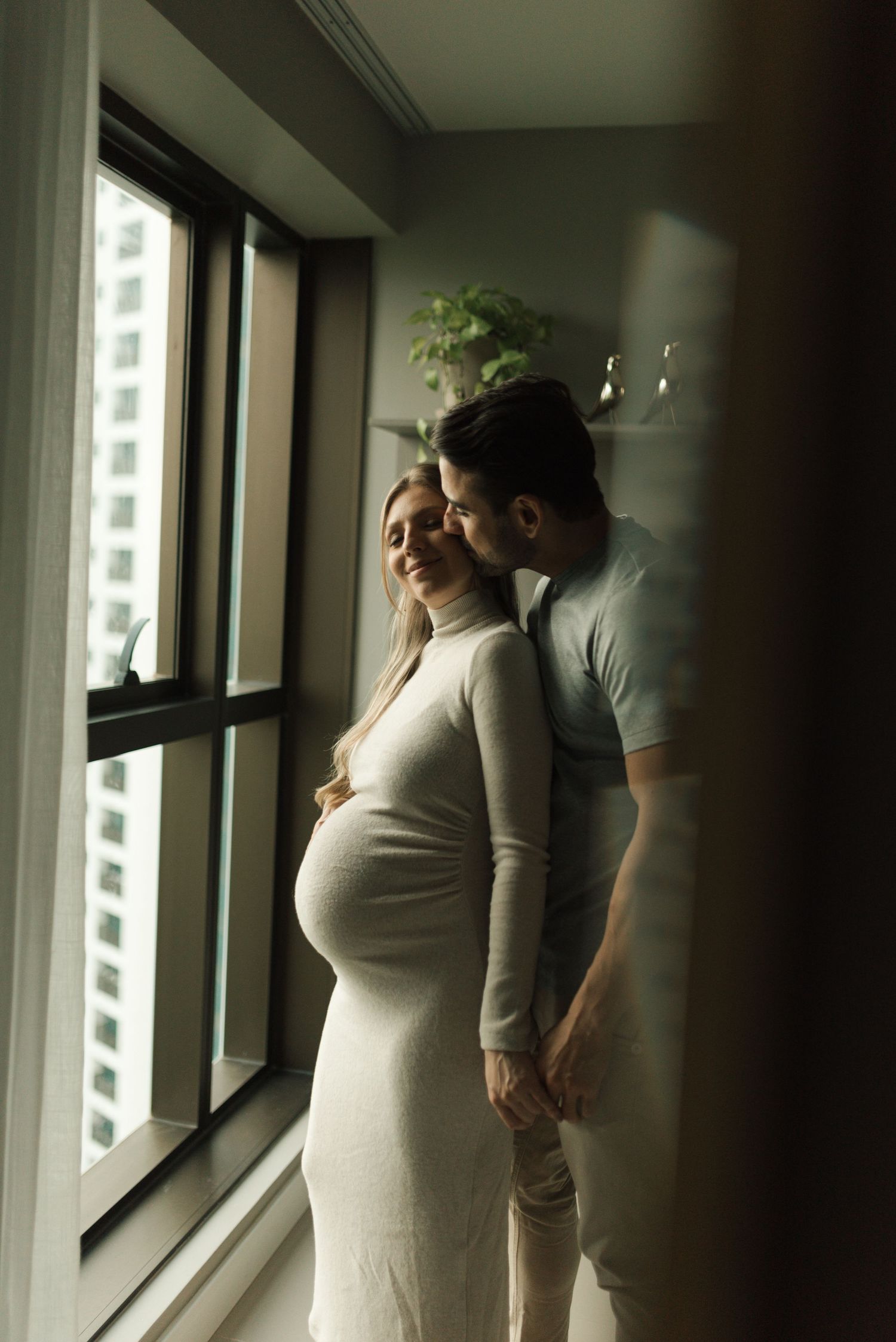Intimate maternity photo of couple by window in white dress shows tender moment and natural lighting.