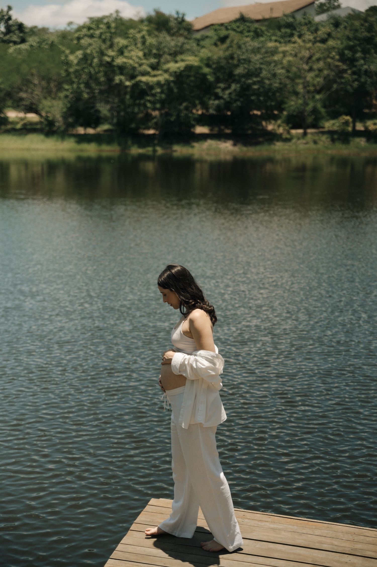 A person in white clothing sits thoughtfully on a wooden dock overlooking a serene lake surrounded by trees.