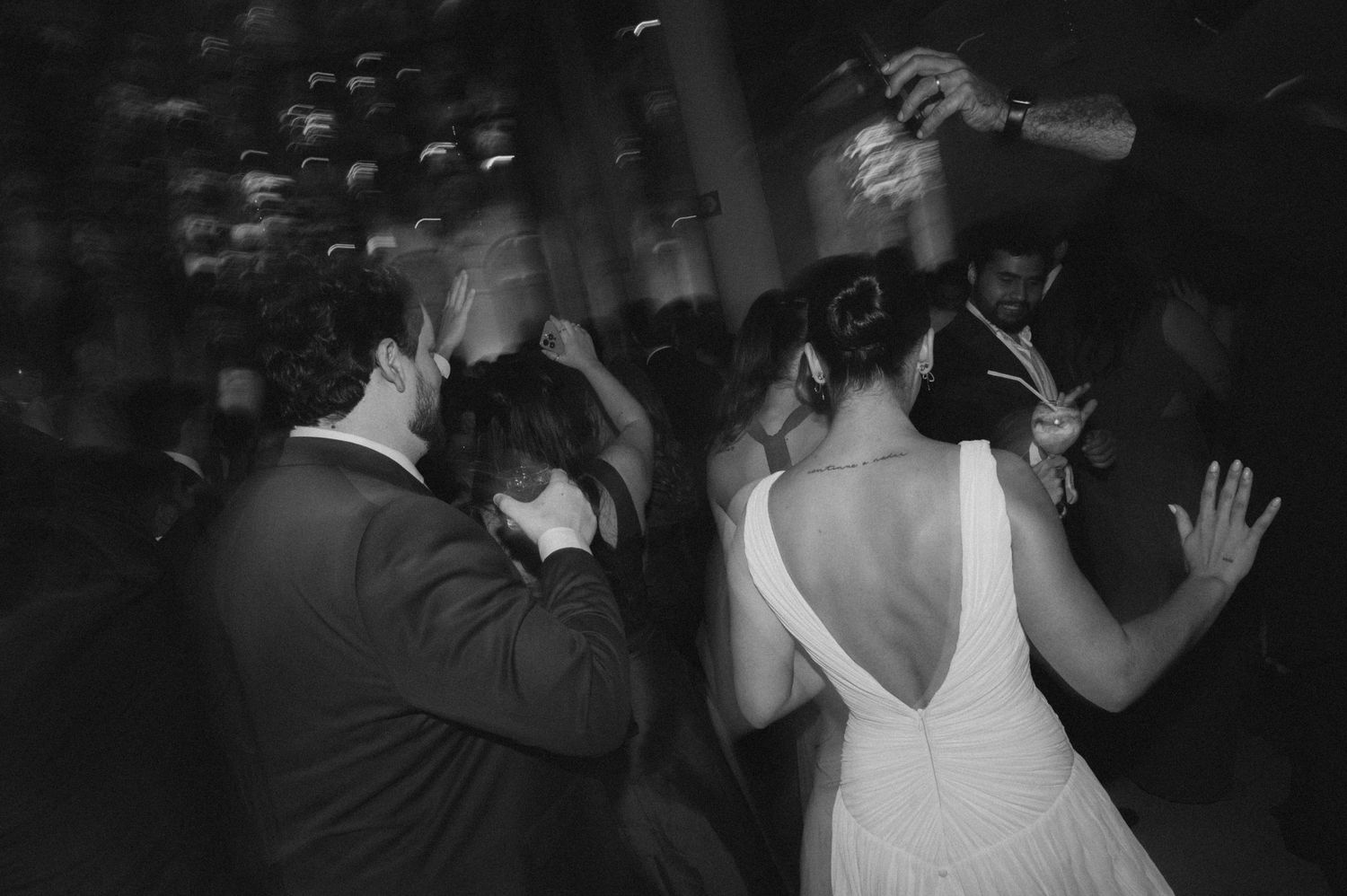 Wedding guests dancing and celebrating in a dimly lit reception venue with arms raised.