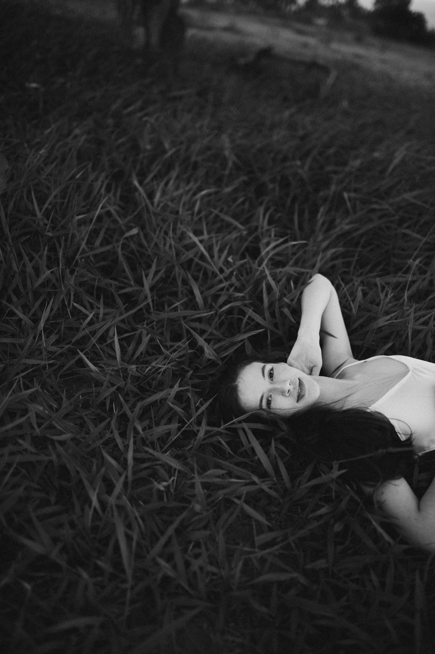 A person in white lies in tall grass while smiling at the camera in black and white.