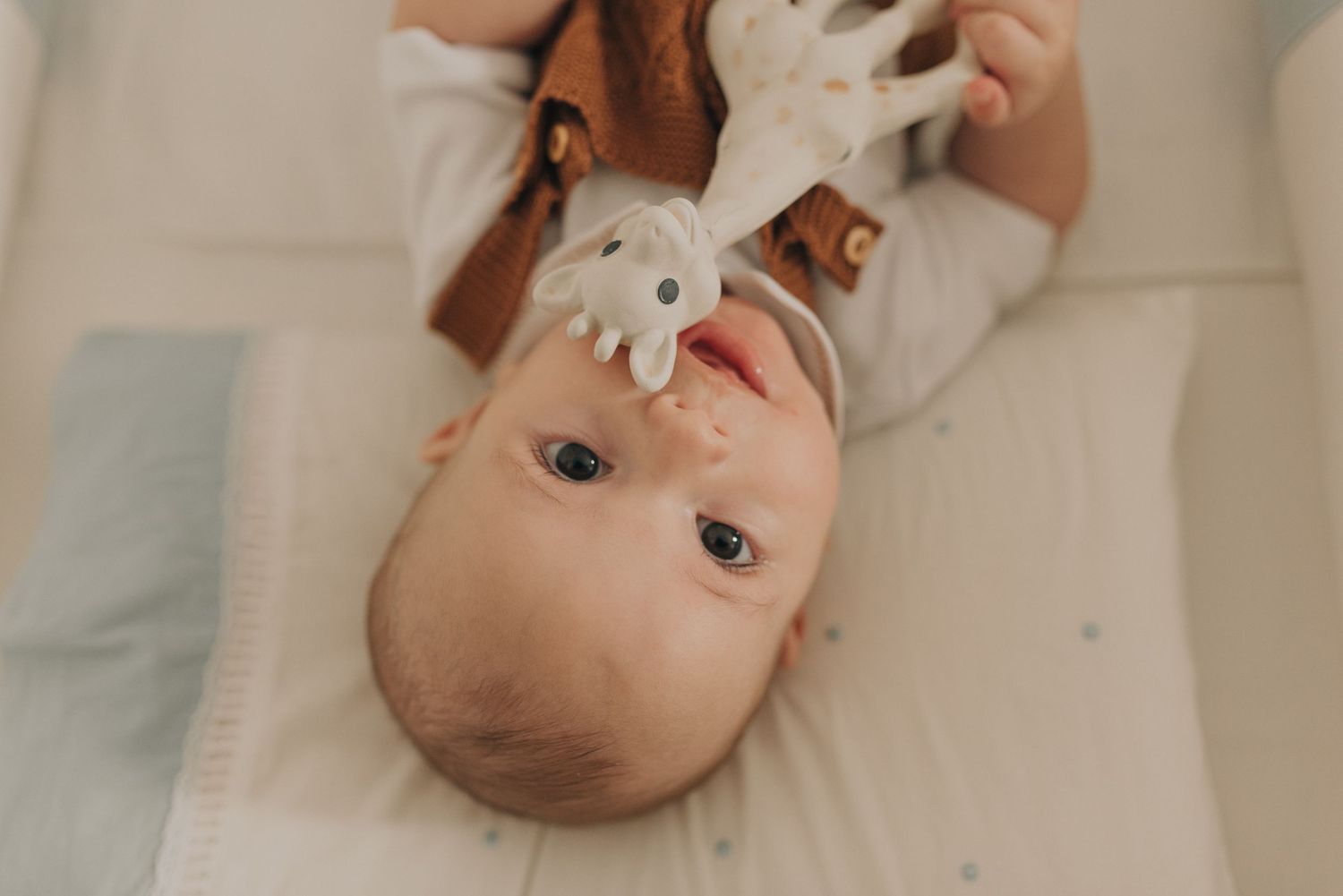 An adorable infant in white clothing lies upside down on a light colored surface looking at the camera.