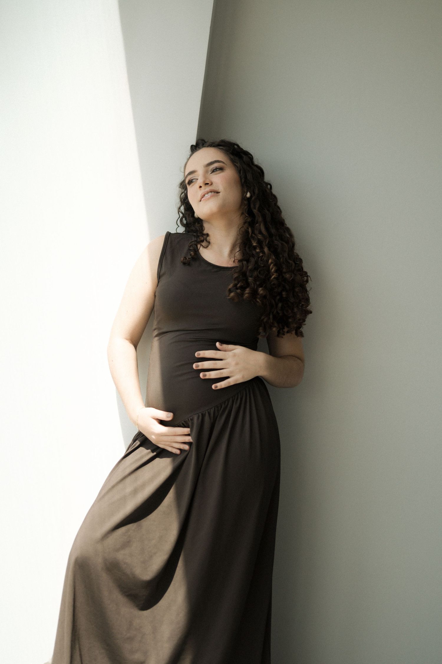 Pregnant woman in dark brown maxi dress leaning against wall, cradling baby bump with curly hair flowing.