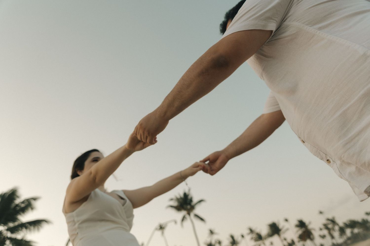 A couple holding hands in white outfits with palm trees silhouetted against a sunset sky.