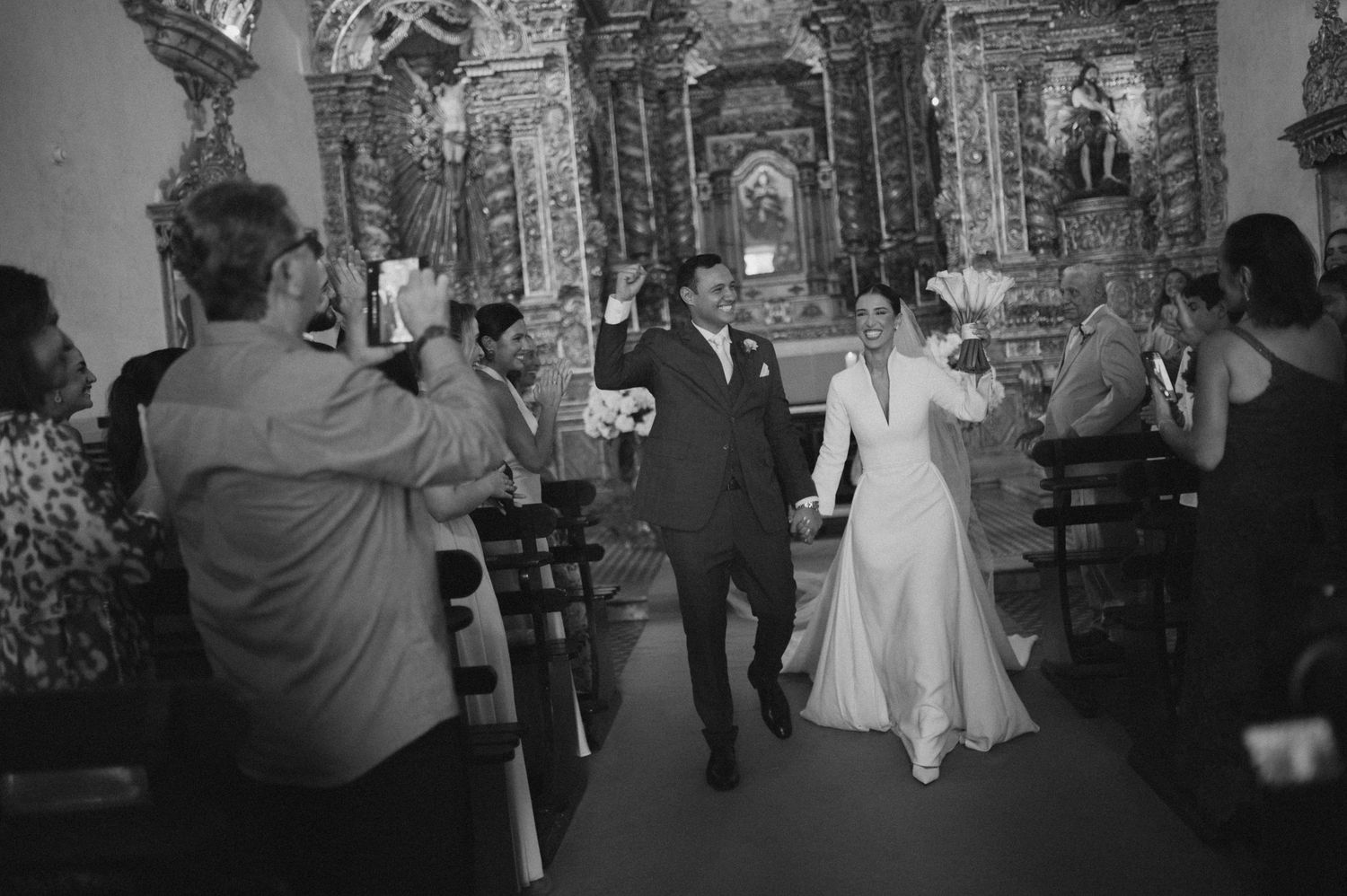 Bride and groom walk down the aisle of an ornate church while guests celebrate their wedding ceremony.