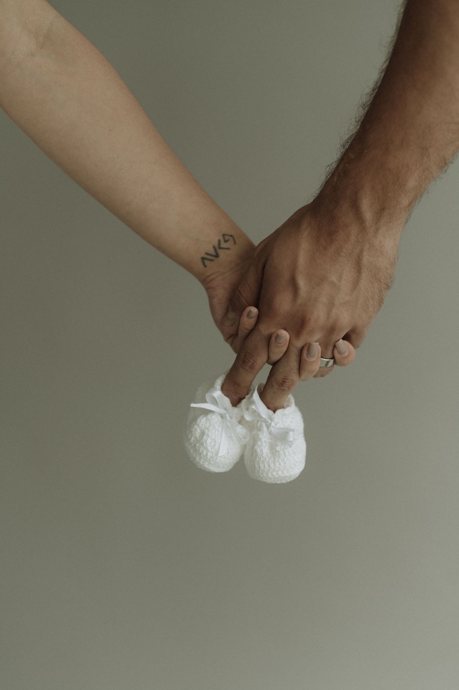 Two hands holding tiny white baby shoes with a small matching tattoo visible on one wrist.