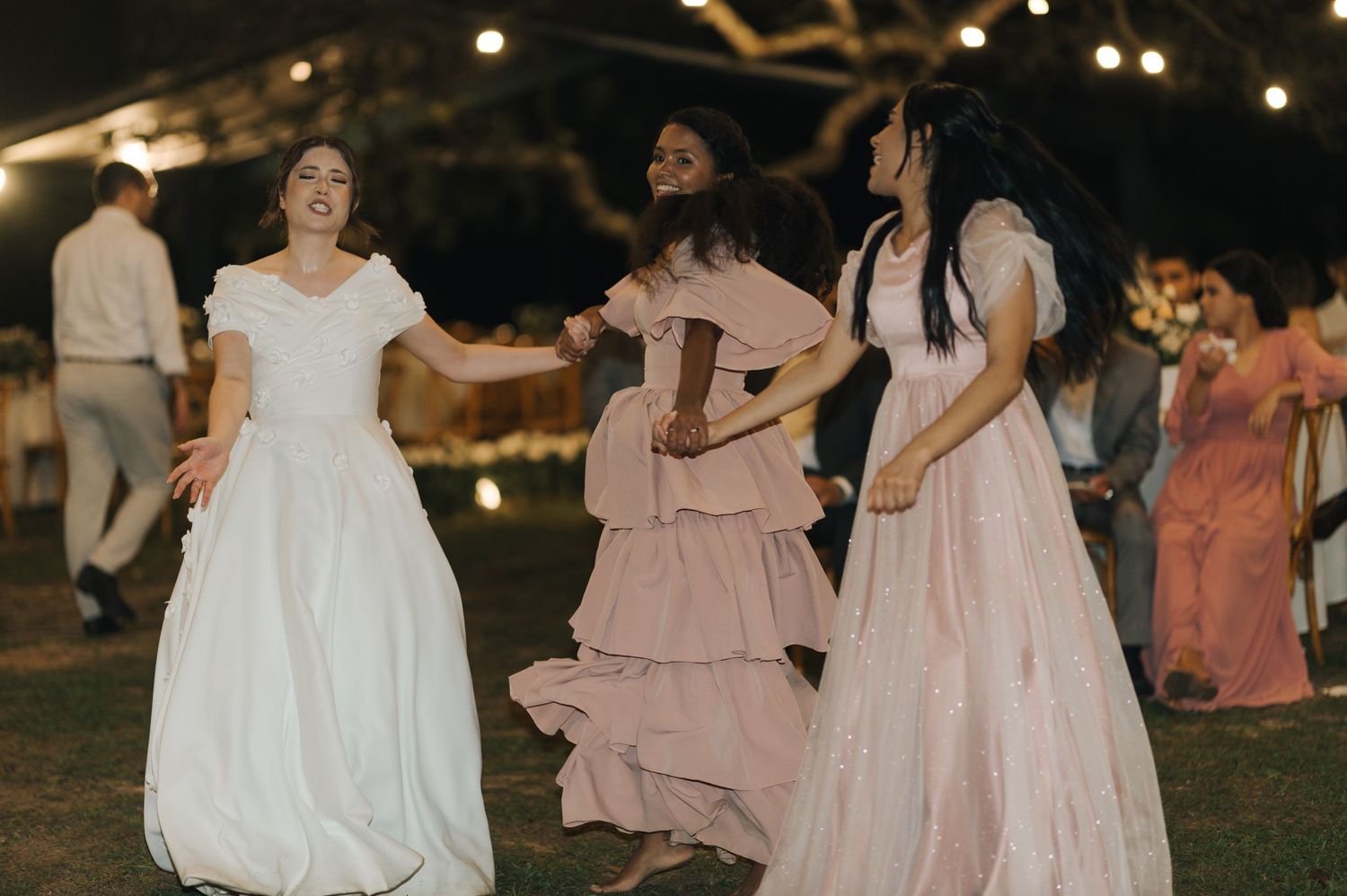 Wedding guests dancing together in flowing dresses under evening lights at an outdoor reception.