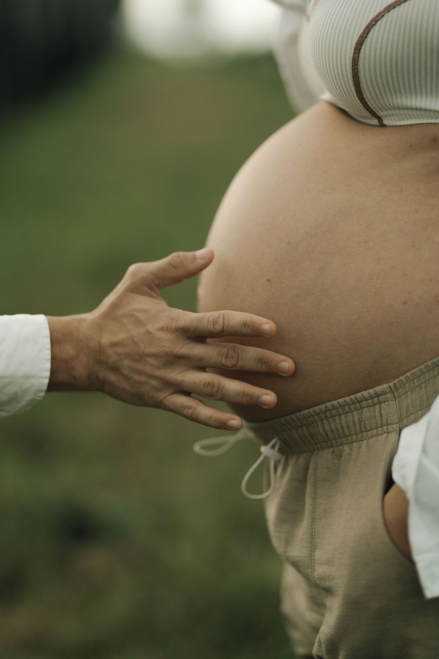 A gentle hand rests on a pregnant belly in a tender maternity photo taken outdoors with soft natural lighting.