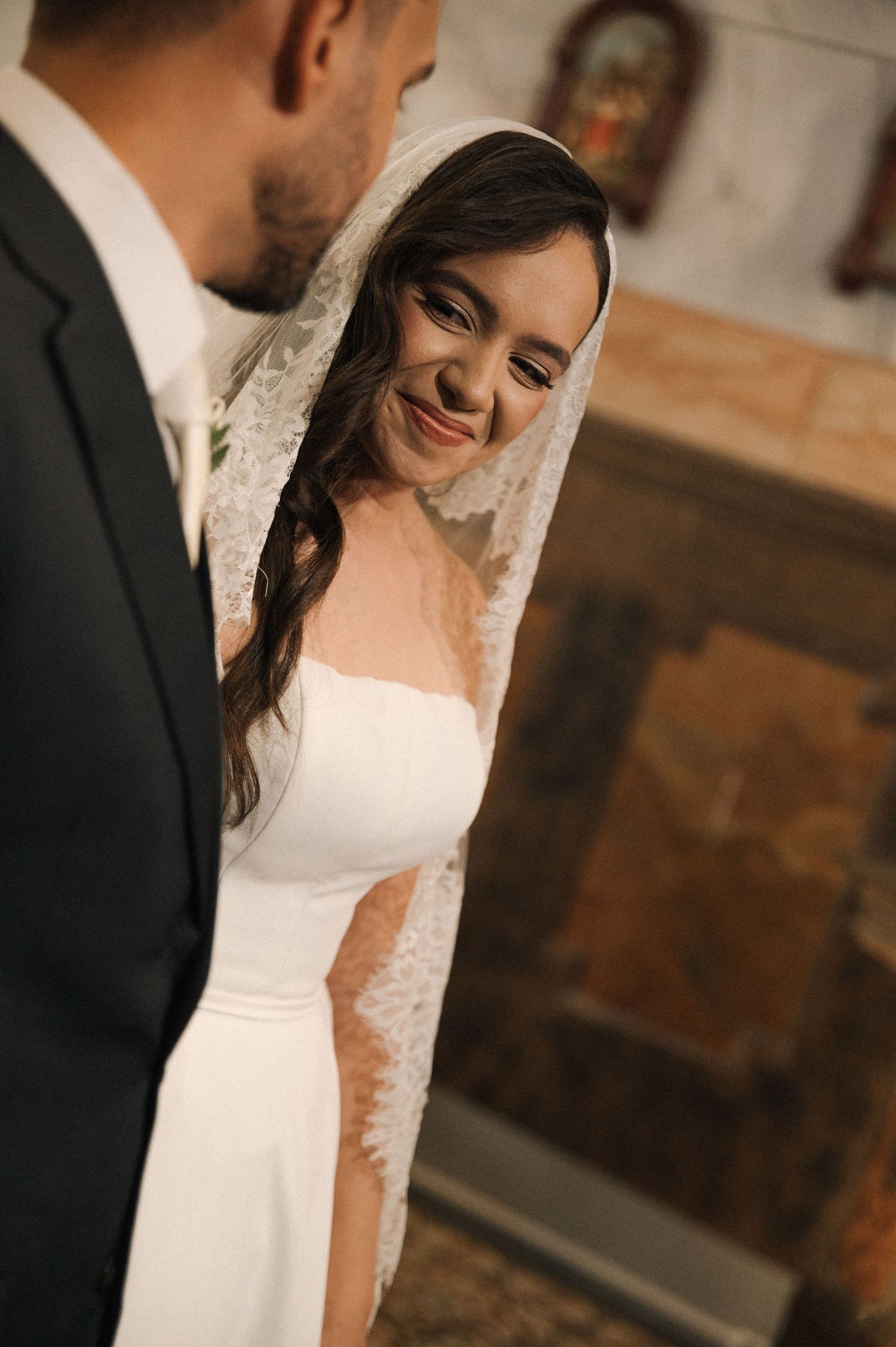 Bride in white gown and lace veil smiles at groom during wedding ceremony in a church.
