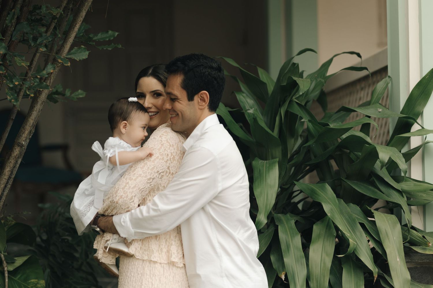 A tender moment shared between parent and baby near lush green indoor plants by a window.