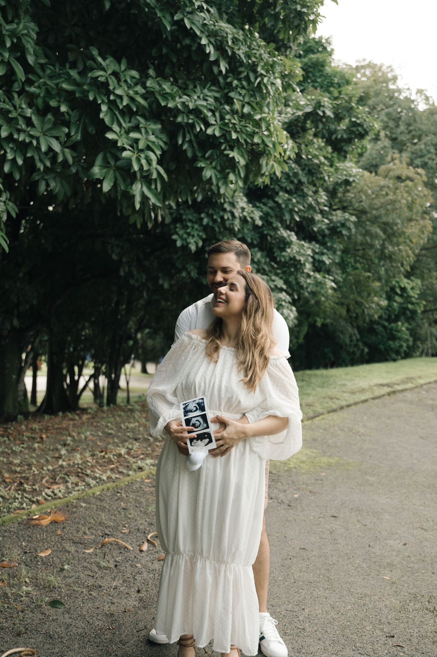 Happy couple in a park holding ultrasound photos, woman in white dress laughing, pregnancy announcement outdoors.