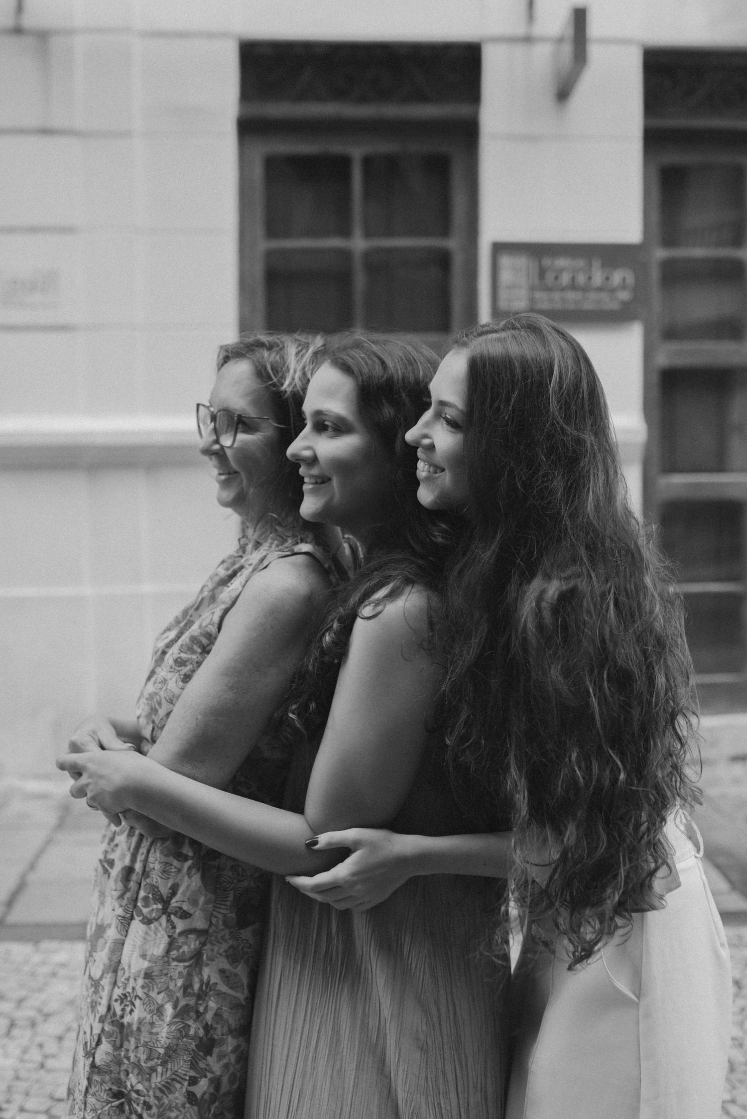 Three women standing together in black and white portrait against urban backdrop.