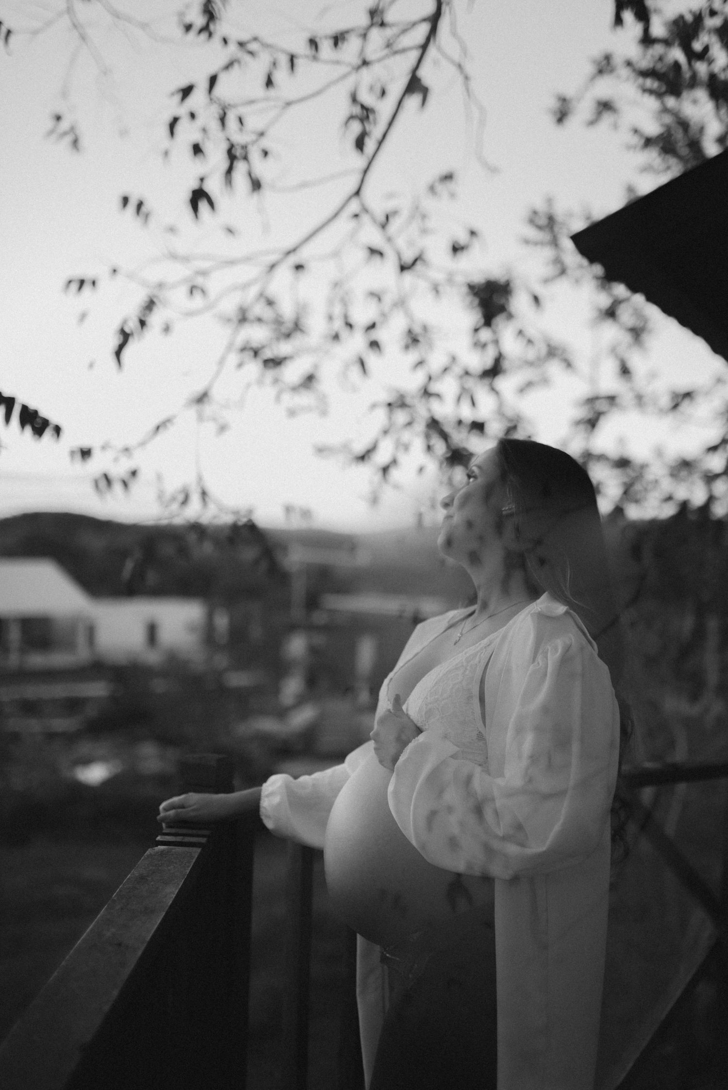 Silhouetted maternity portrait against sunset sky with woman in white dress on balcony.