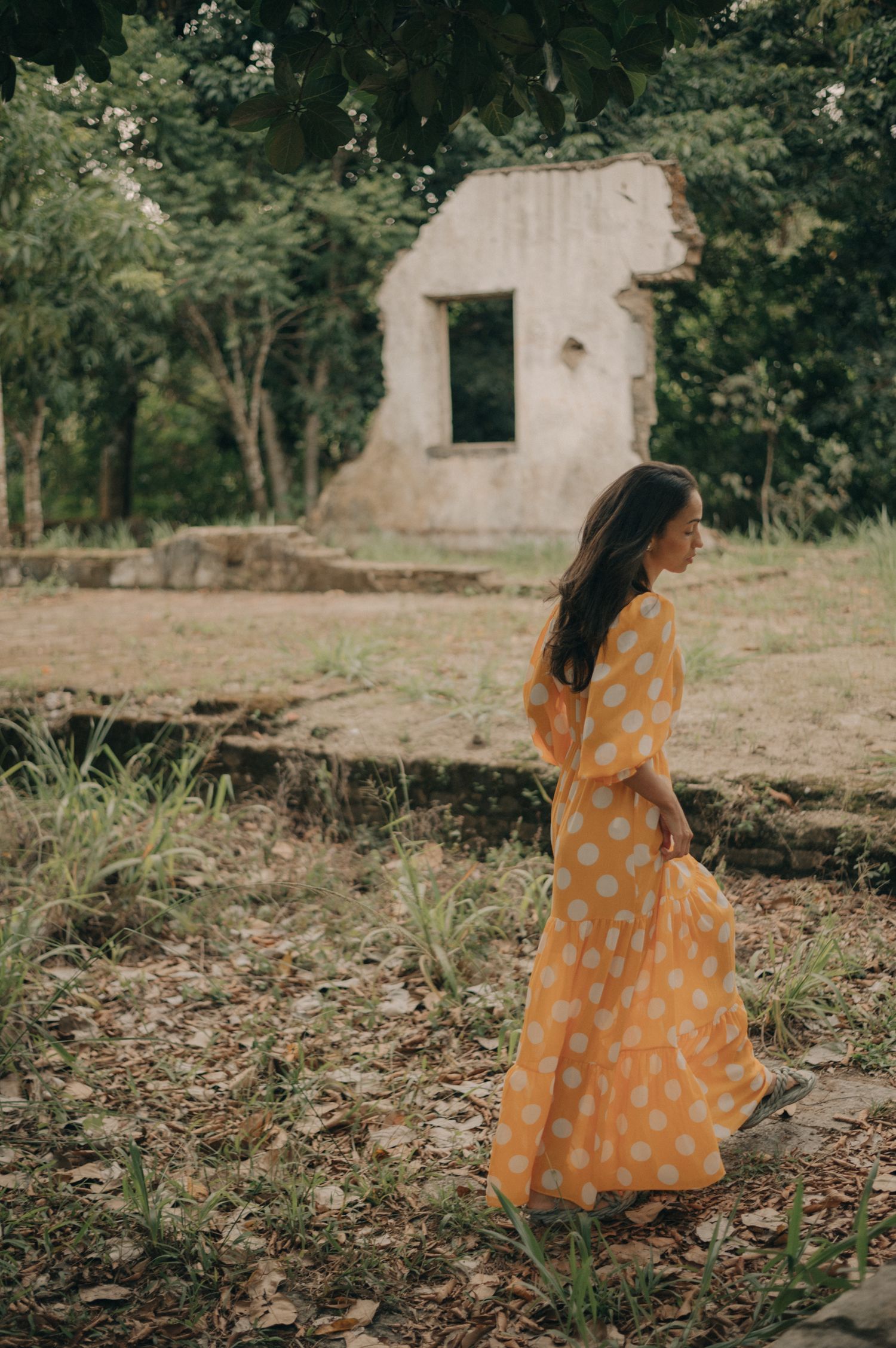 A person in a flowing polka dot dress walks near an old stone structure surrounded by greenery.