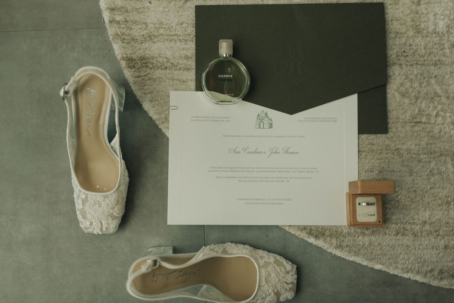 Wedding invitation suite with elegant calligraphy, white ballet flats, and perfume bottle arranged on textured gray surface.