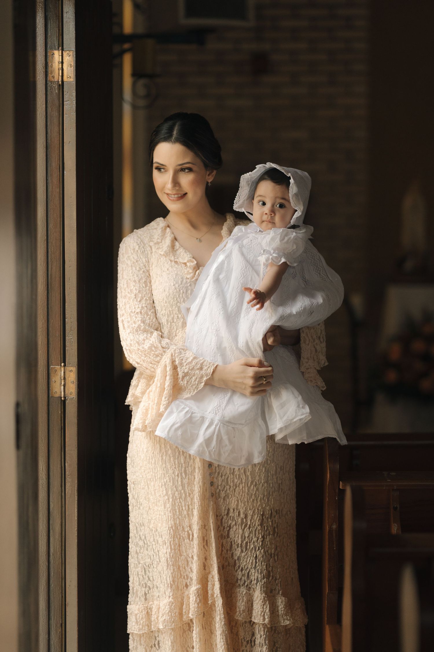 A person in a cream lace dress holds a baby in white christening gown by a window in warm natural light.