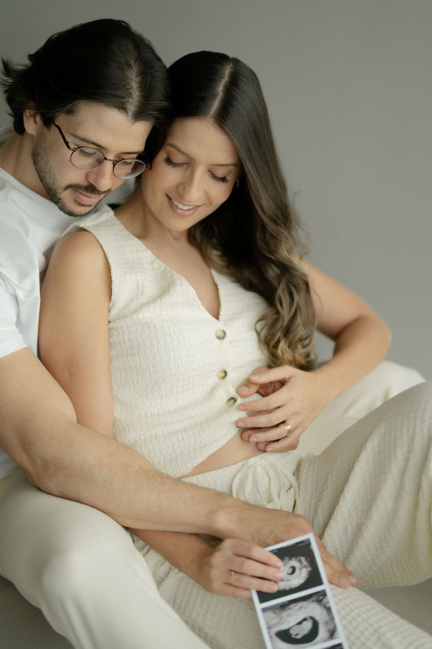 Couple in white clothing looks lovingly at ultrasound photo while embracing during pregnancy photoshoot.
