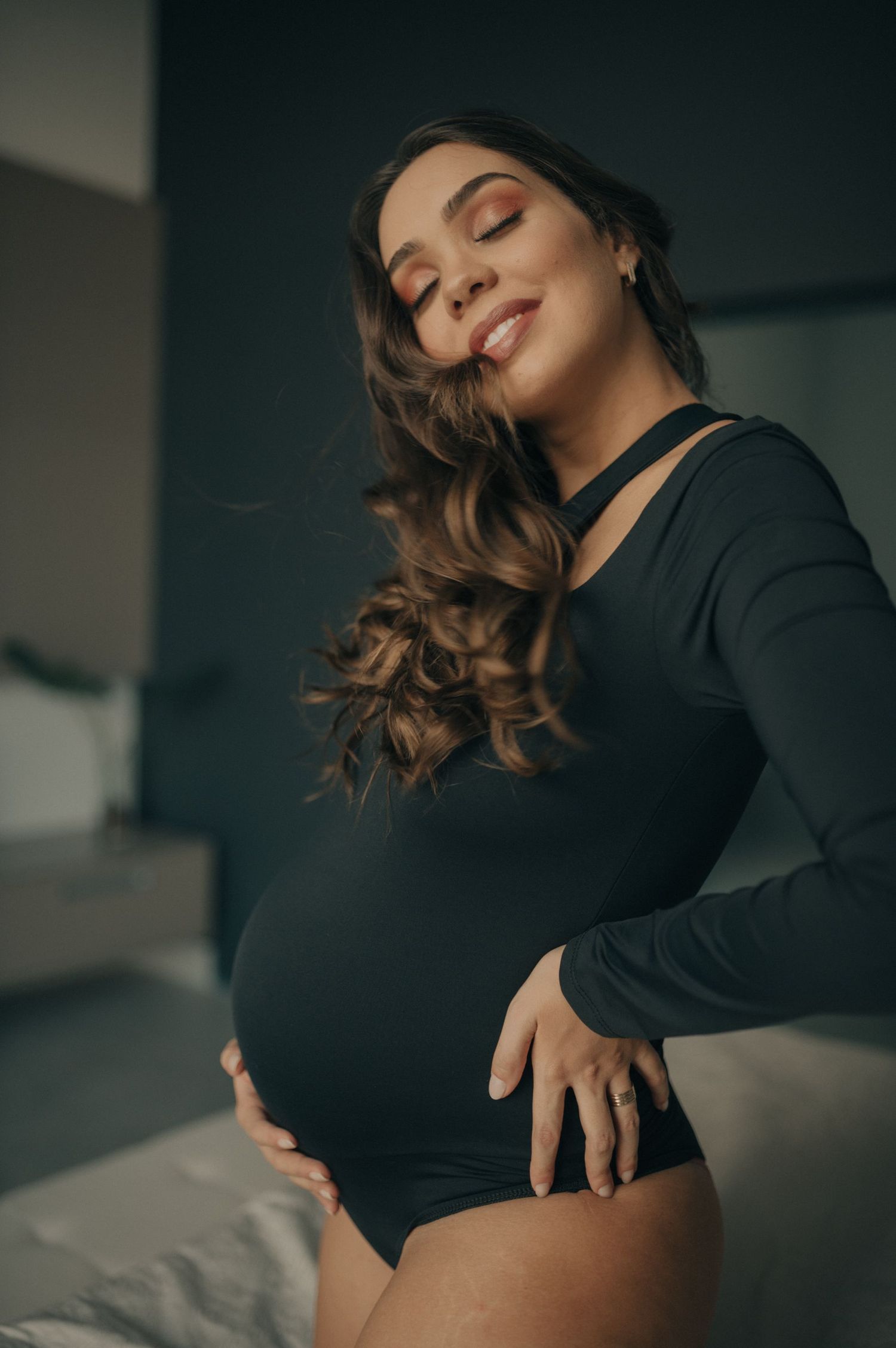 Moody maternity portrait in black bodysuit against dark interior backdrop.
