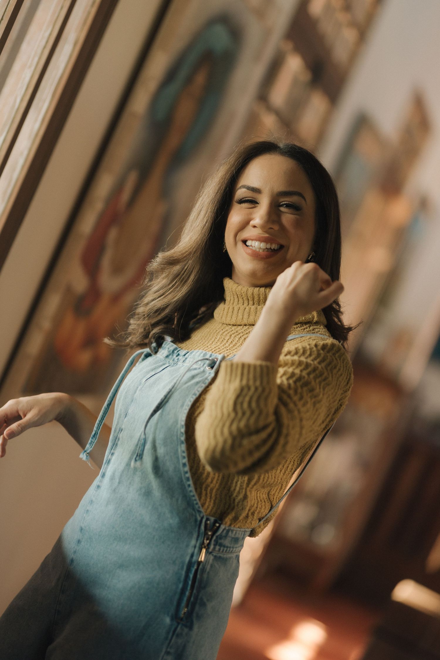 Candid indoor portrait of someone wearing denim overalls and a mustard turtleneck sweater.