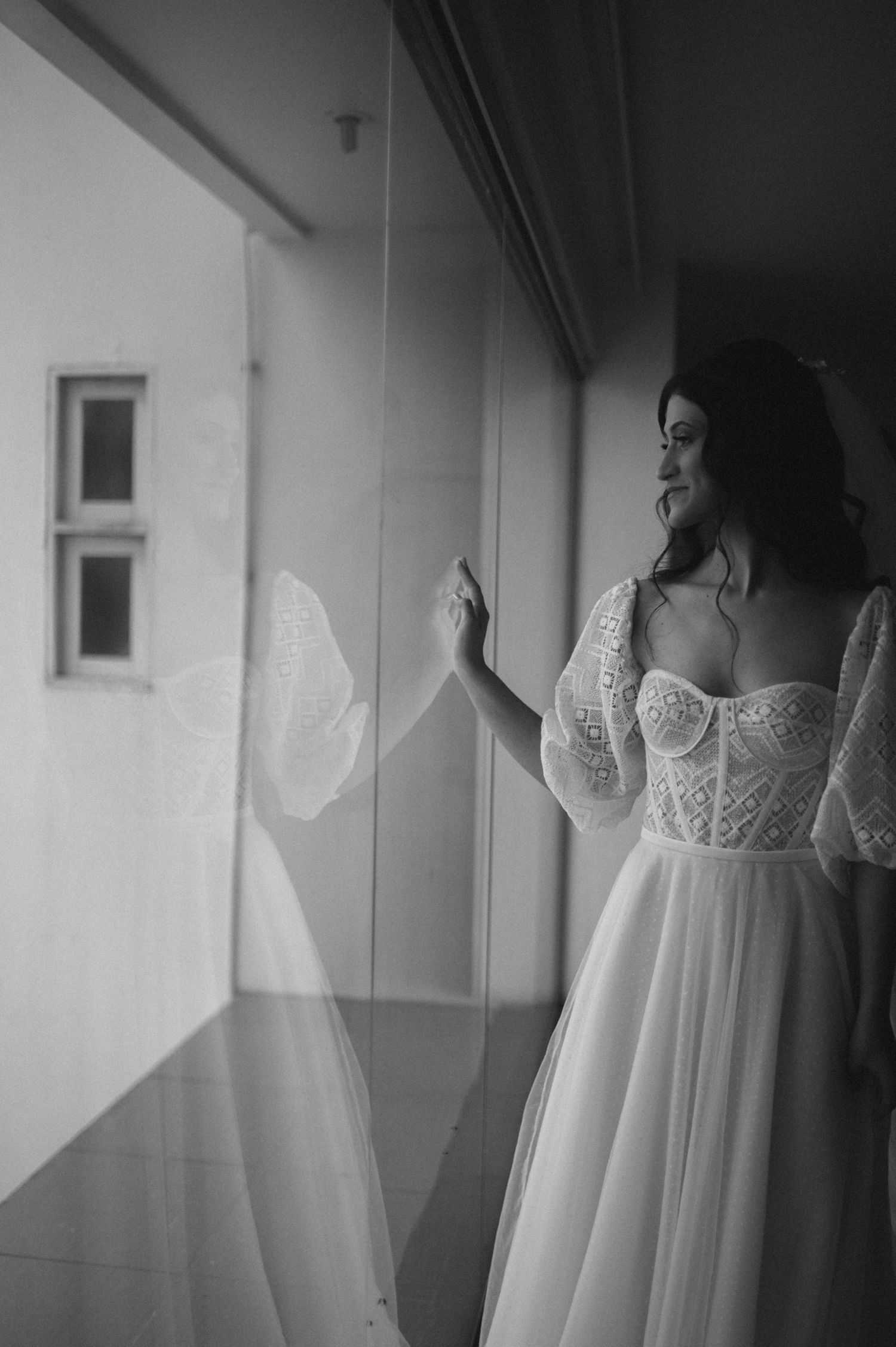 Black and white photo of a bride in a corset dress looking out a window.