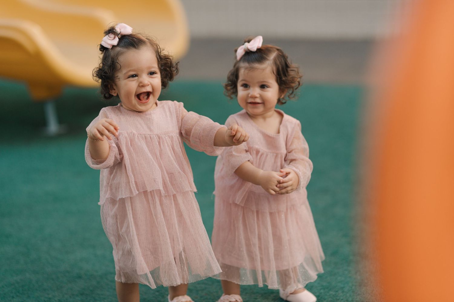 Two toddlers in matching pink dresses play and laugh together on green turf.