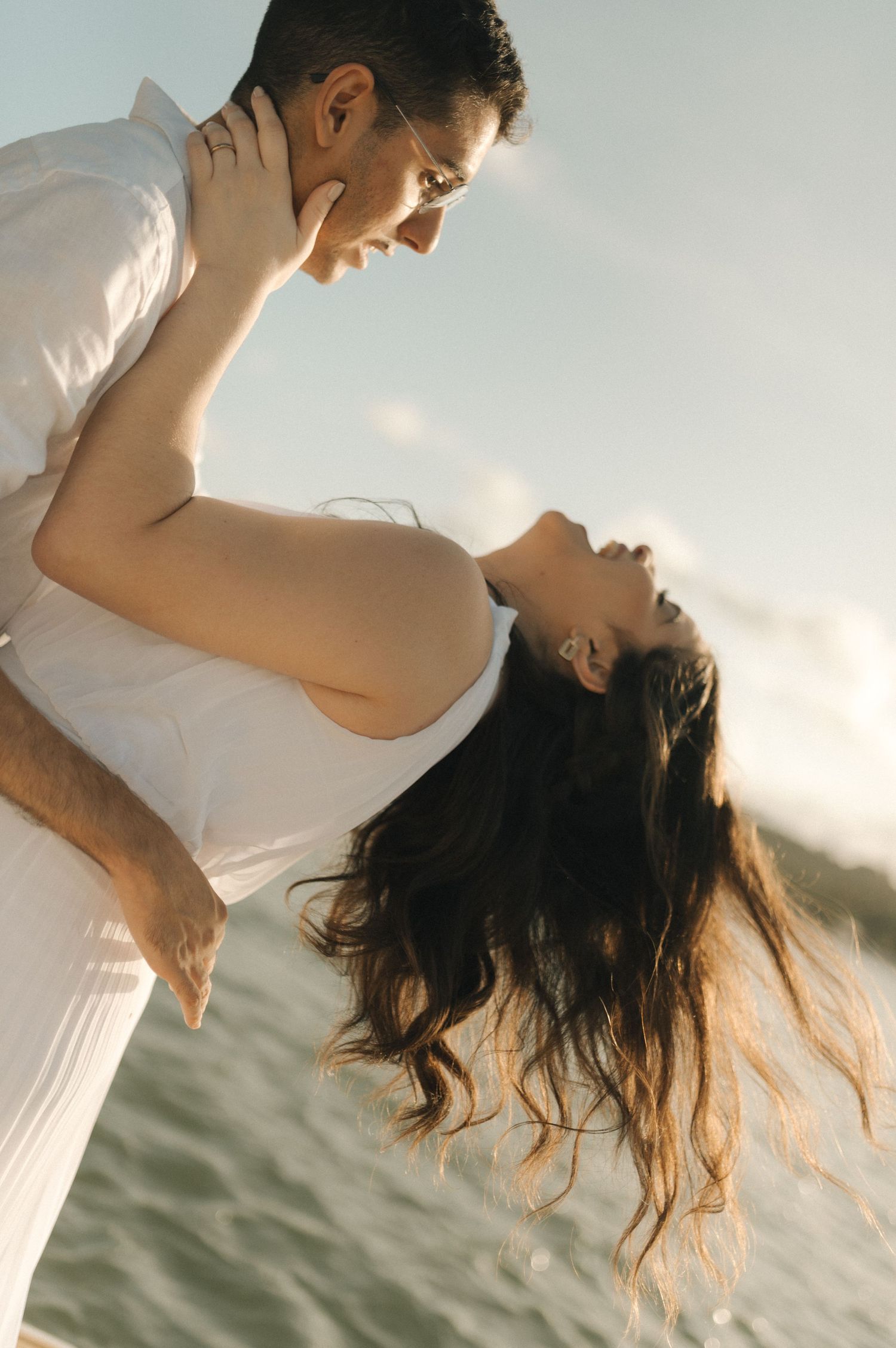 Couple in white clothes sharing an intimate moment at sunset with dramatic lighting and ocean backdrop.