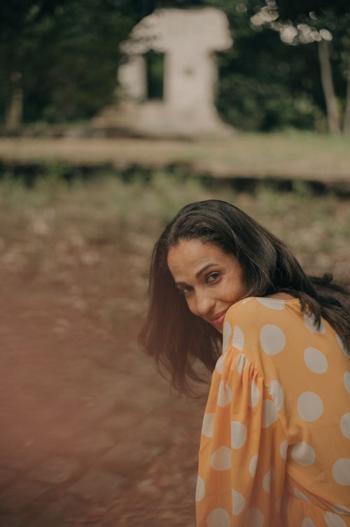 A person in a polka dot yellow dress stands near a rustic stone structure in a natural outdoor setting.
