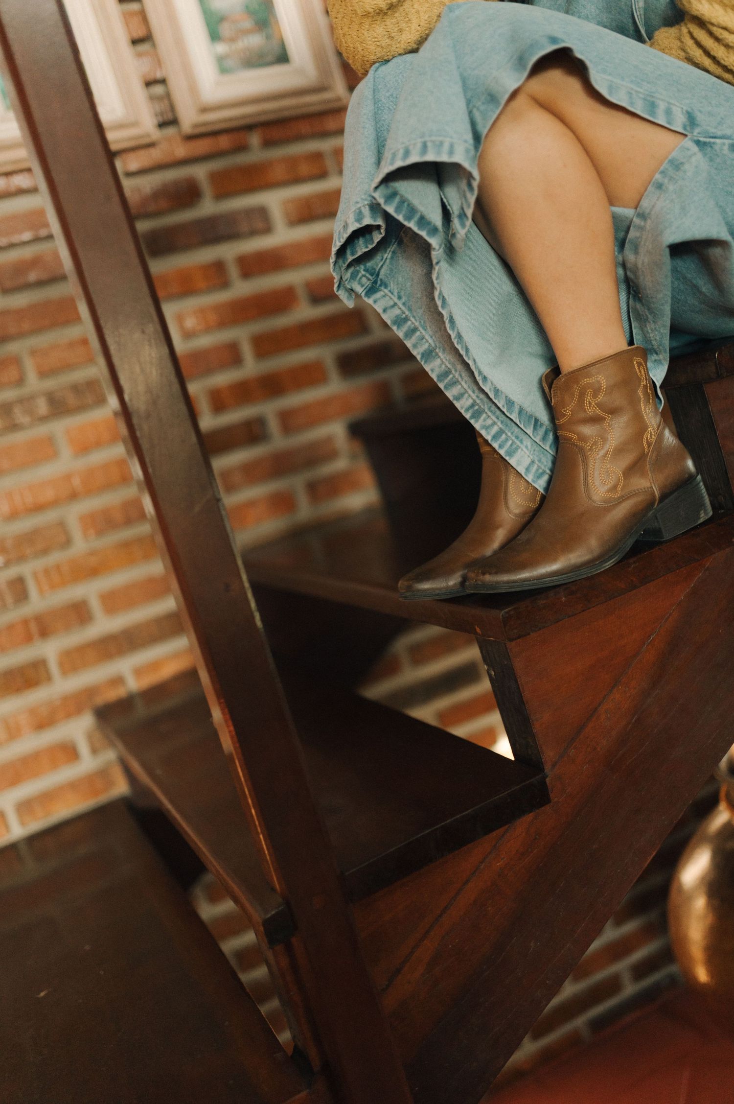 Brown leather cowboy boots and denim outfit visible on wooden stairs against brick wall.