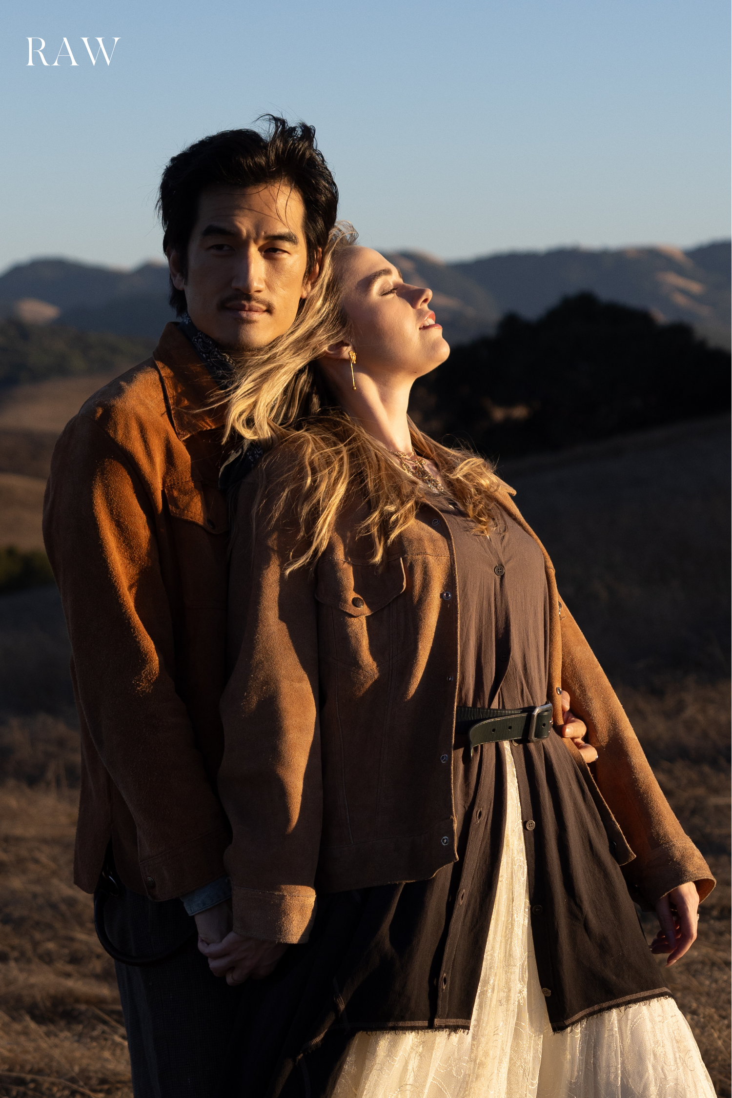 Couple in vintage-inspired clothing pose together against golden hills during sunset.