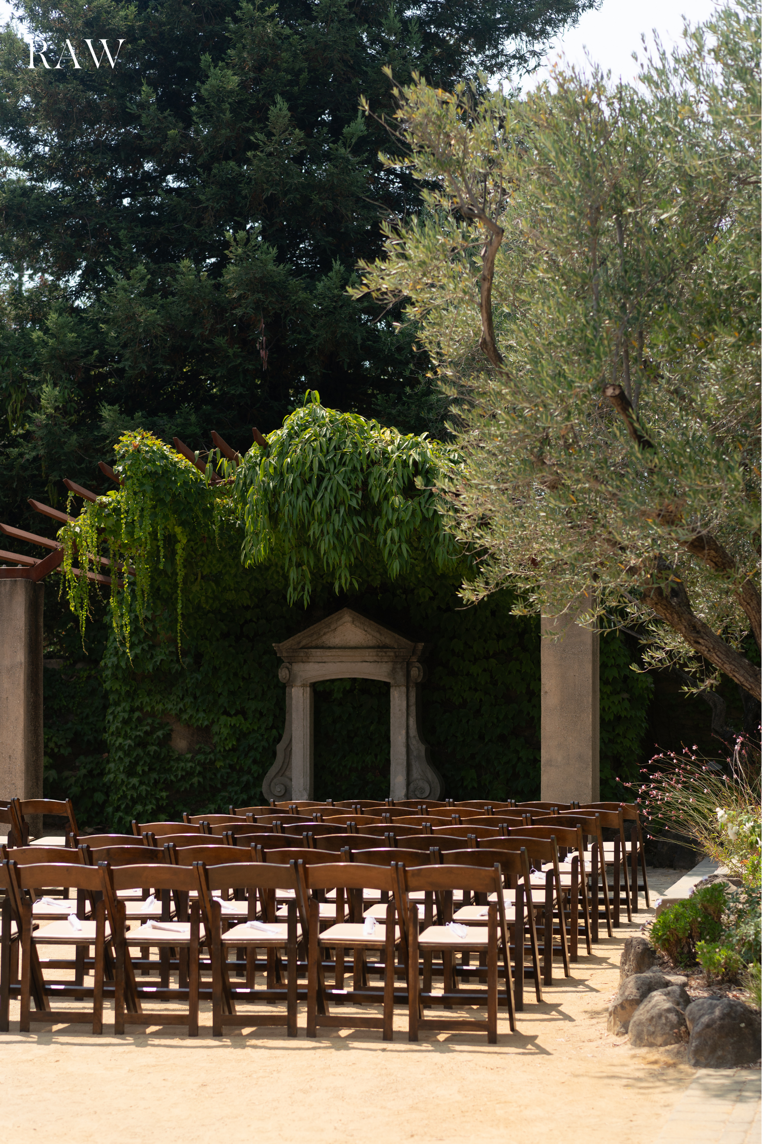 Rustic wooden wedding chairs arranged in rows under a canopy of trees.