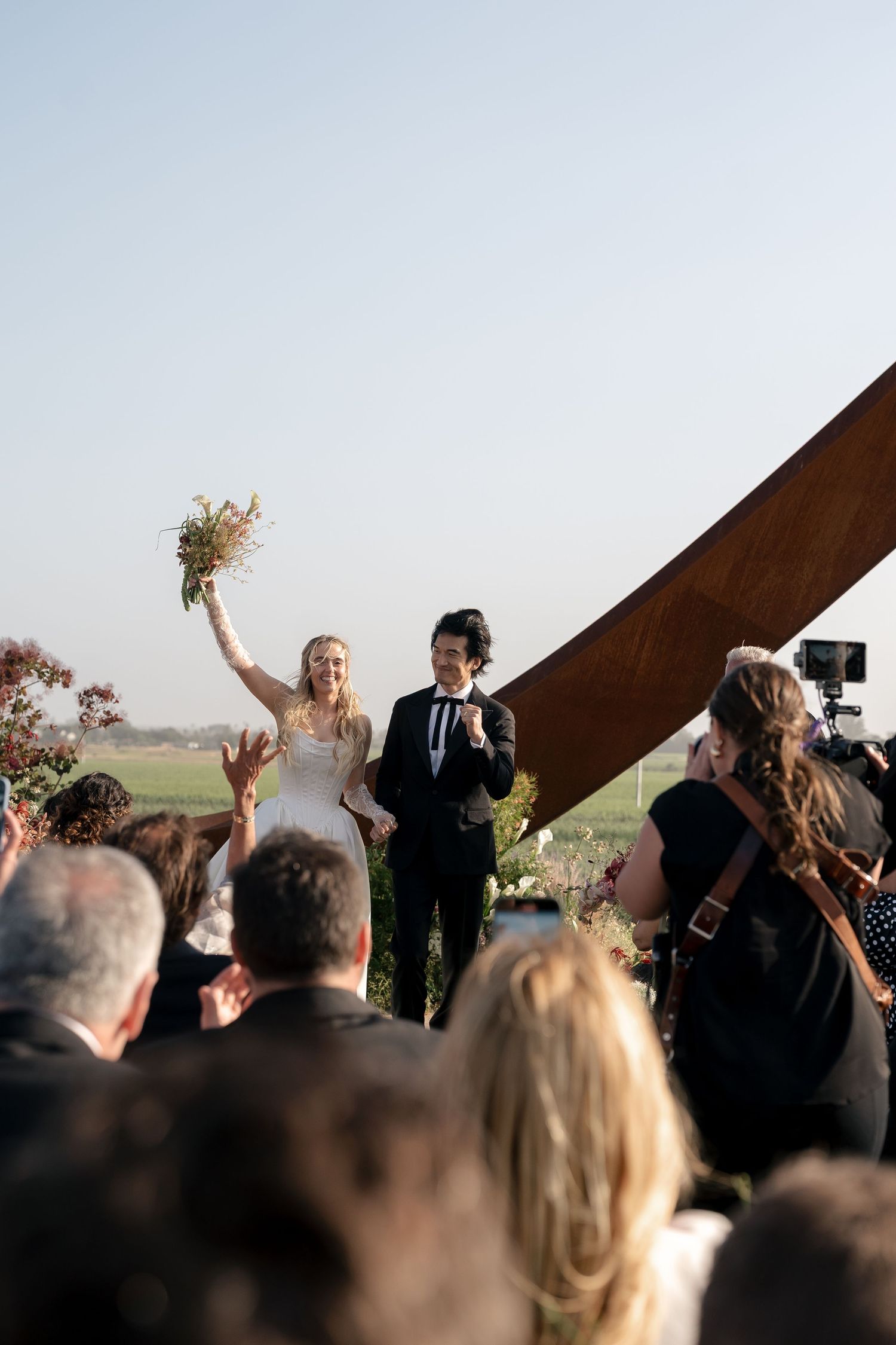 Wedding guests celebrate outdoors with raised arms against a dramatic architectural backdrop.